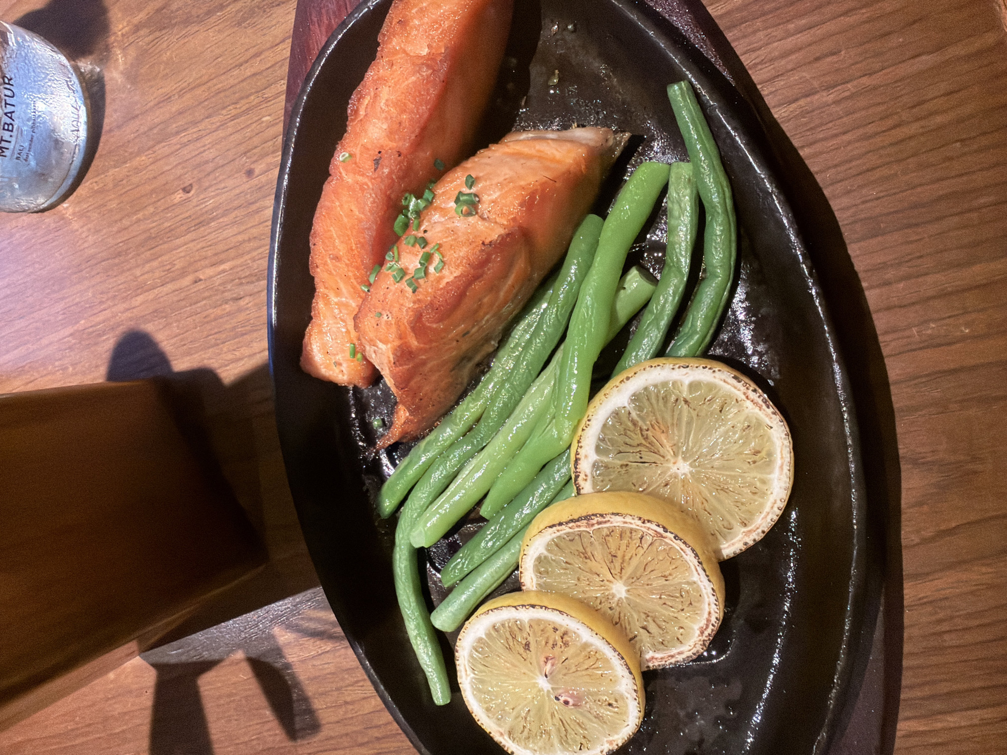 A close-up, eye-level shot features a sizzling hot plate of food with two salmon fillets, a side of vibrant green beans, and three slices of grilled lemon. The salmon, cooked to a perfect medium, is garnished with finely chopped chives. The hot plate rests on a polished wooden table, where a glass of amber liquid casts a subtle shadow. In the background, a partially visible bottle of mineral water with "MT. Batur" branding suggests a refined dining experience, possibly at a restaurant in Central Jakarta, Indonesia, known for its upscale establishments. The lighting is warm and inviting, highlighting the textures and colors of the food.