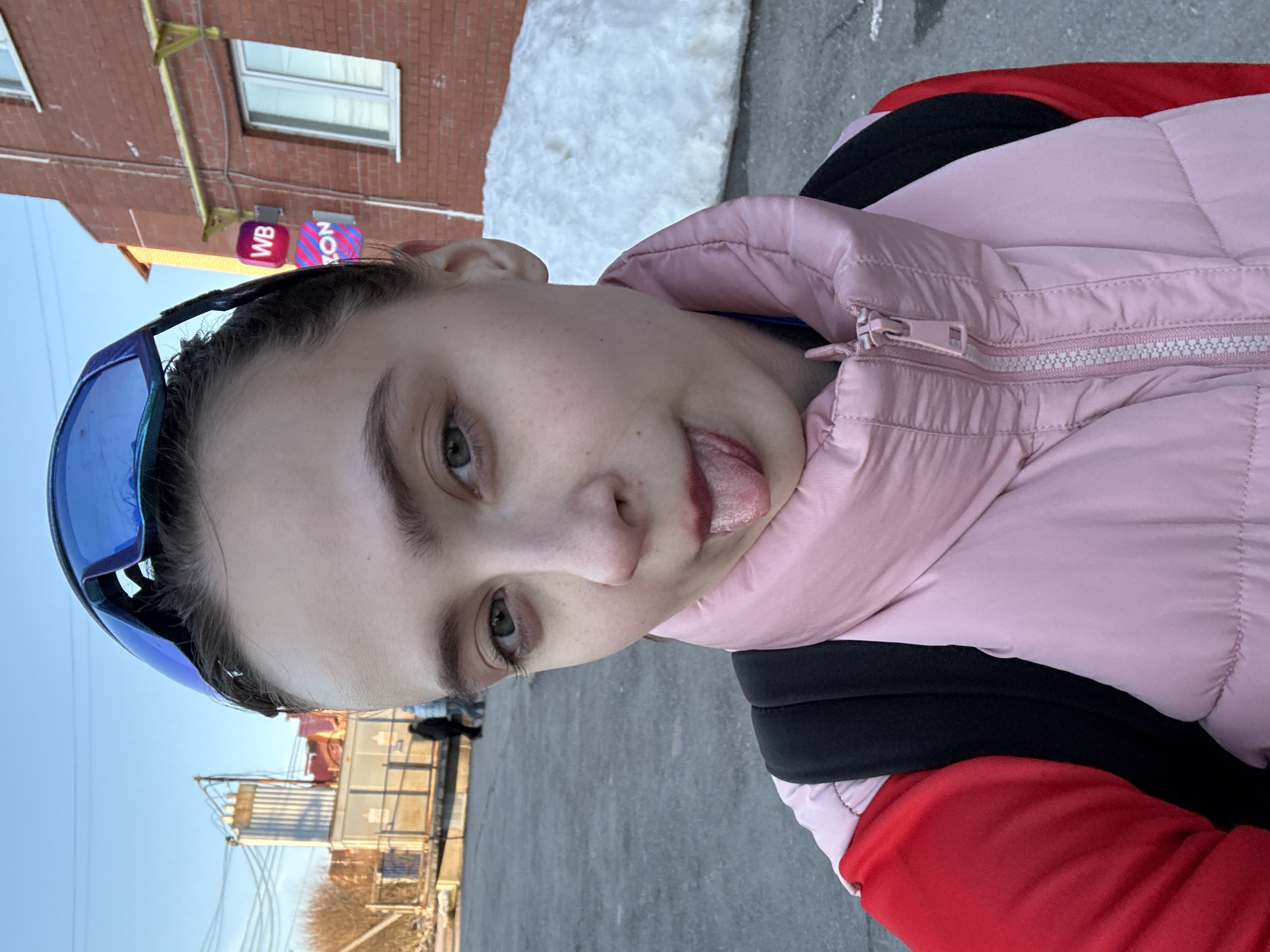 A photograph shows a person, presumably a young woman, with short dark hair and fair skin, sticking out her tongue. She is wearing blue-tinted glasses pushed up on her head and a light pink puffer jacket with a black backpack underneath. The background includes a building with a brick facade and white-framed windows. There's a small sign featuring "WB" in pink and purple. A patch of snow is visible near the building. The ground is grey pavement. In the distance, there are industrial structures and bare trees against a blue sky. The location is likely Berezniki, Russia, based on the "WB" sign, which could indicate a local business or landmark associated with that location.