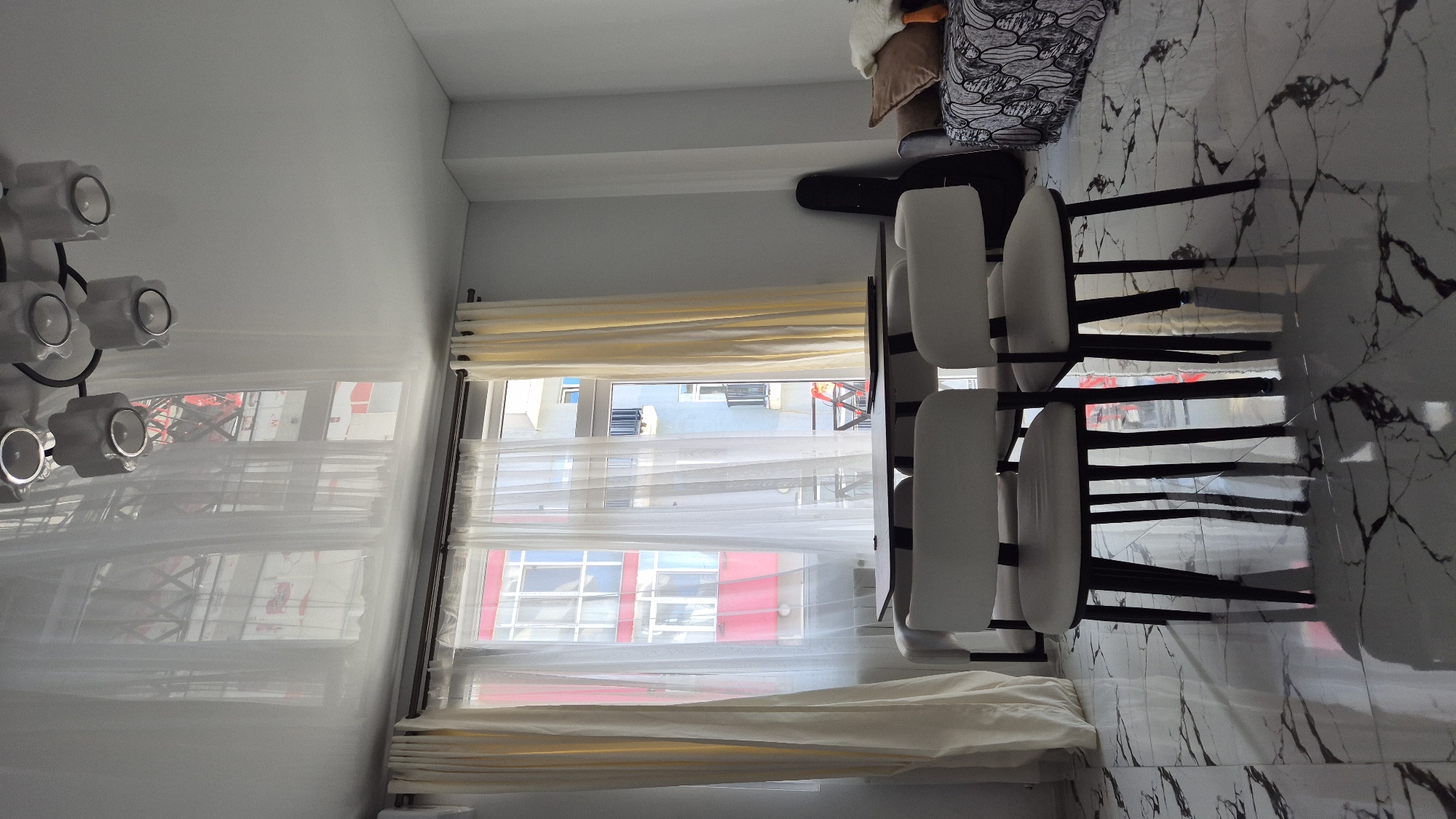 The image shows the interior of a room, possibly an apartment or hotel room, in Batumi, Georgia. The room has a window with sheer white curtains and heavier cream-colored curtains along the top and bottom. Outside the window, there are buildings in a blue, red, and white color scheme, indicating an urban environment. The room is furnished with several chairs with white seats and black frames, stacked together on a marble patterned floor. In the corner, there is a sofa or bed with various pillows. A unique light fixture with multiple circular lamps is mounted on the wall. There are no people visible in the image.