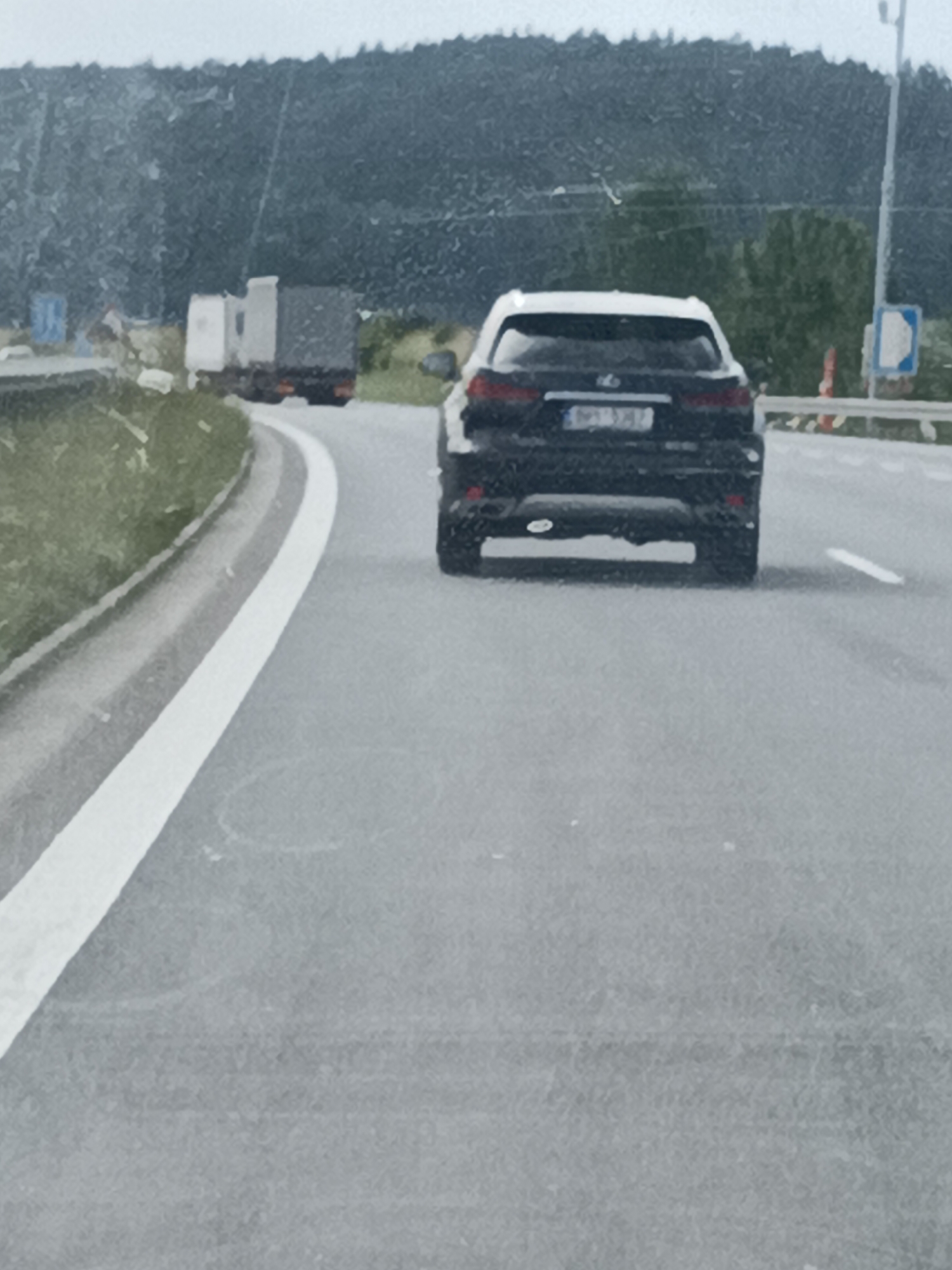 This media file captures a rear view of a black Lexus SUV driving on a paved road. The SUV is positioned in the right lane, moving away from the camera. Its license plate is visible and reads "BP1 1347". In the background, a large truck with a light-colored cab and a dark cargo area is also visible, driving ahead and to the left of the SUV. The road curves to the left, bordered by a grassy embankment on the left and a barrier with traffic signs on the right. The sky appears overcast, suggesting a cloudy or possibly rainy day. The overall scene depicts a moment of road travel, likely on a highway or a major road, with a forested hill in the far background. The lighting is diffused, typical of an overcast sky. The image quality is somewhat grainy, suggesting it was taken with a mobile device or under low-light conditions. There are no people clearly visible in the image.