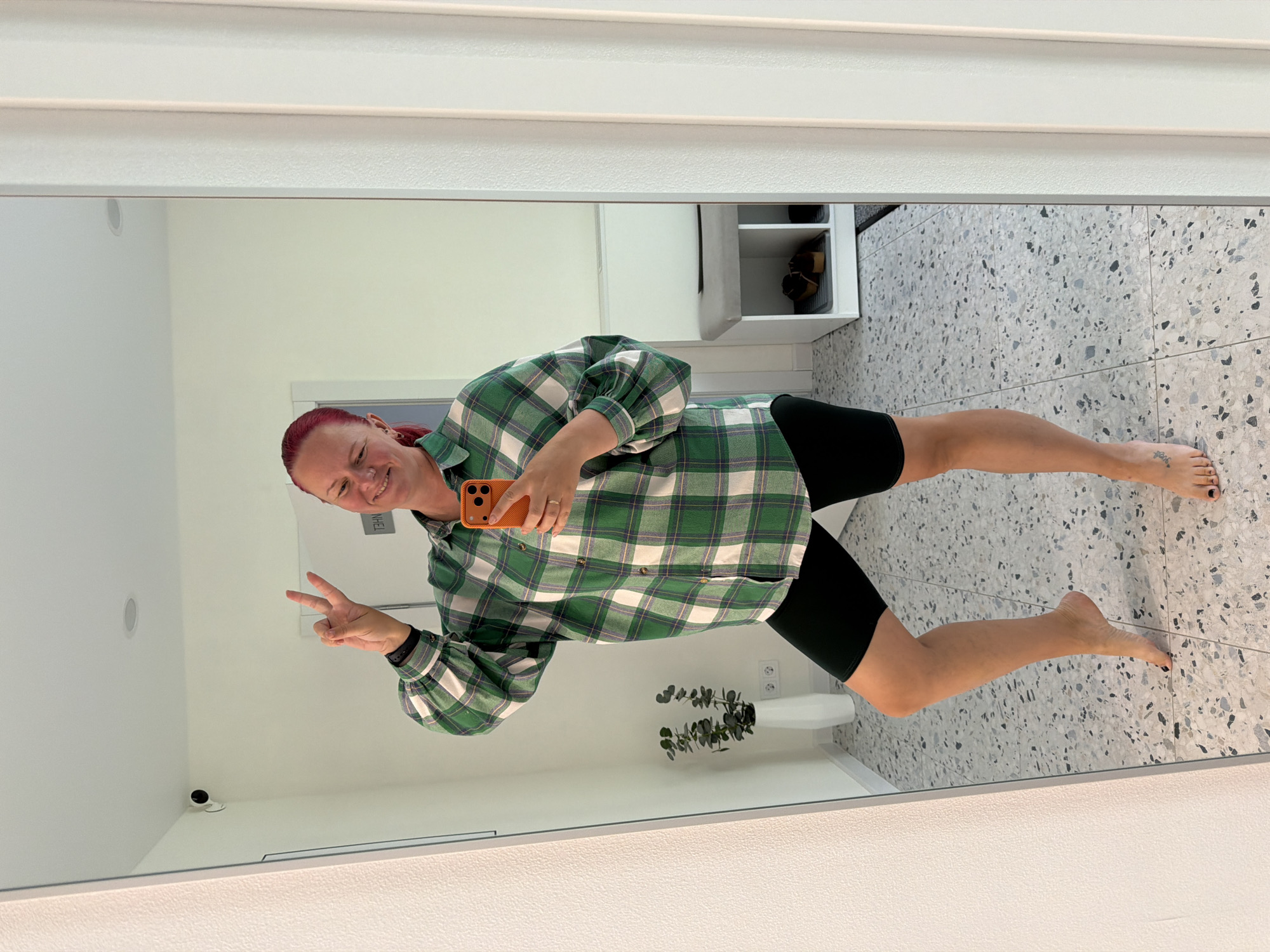 An adult female with red hair, wearing a green, white, and dark blue plaid long-sleeved shirt and black cycling shorts, stands barefoot, posing for a mirror selfie. She holds an orange smartphone in her left hand, smiles, and makes a peace sign (V-sign) with her right hand. A dark fitness tracker is visible on her right wrist, and a ring is on her left ring finger. A small tattoo is present on her left foot. The indoor setting features light-colored walls and a light-toned terrazzo-style tiled floor with speckled grey, black, and white aggregate. A white tapered vase containing an artificial plant with green leaves is located on the left side of the reflected room, with two white double electrical outlets on the wall behind it. In the background right, a white shoe storage bench with a light grey cushioned top displays two pairs of shoes within its cubbies. A small, black dome-shaped security camera is mounted high on the wall in the upper left of the reflected space, and a recessed light fixture is visible in the white ceiling. Reversed text is visible on a wall section to the right of the subject's head. This scene is located in the city of Chaikovsky, Russia.