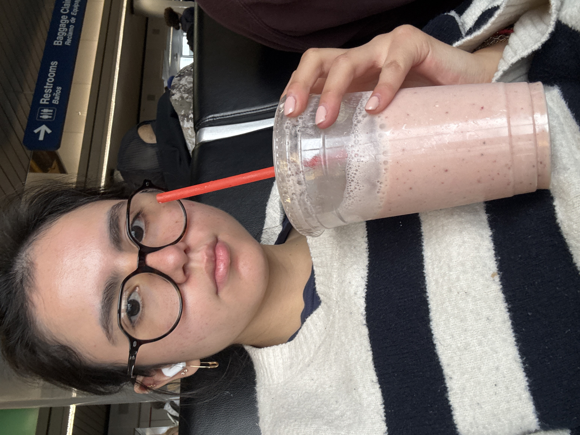 A young woman is seated in an airport terminal. The woman is fair-skinned, with dark hair pulled back, and wearing round glasses. She has multiple earrings in her left ear and is wearing a black and white striped sweater. She is holding a large plastic cup with a pink smoothie and a red straw. The terminal's window is visible, showcasing natural light and a sign for "Restrooms" and "Baggage Claim." Given the airport setting, signage, and general architectural style, the location is likely O'Hare International Airport (ORD) in Chicago, Illinois, USA. Other passengers can be seen sitting in the background.