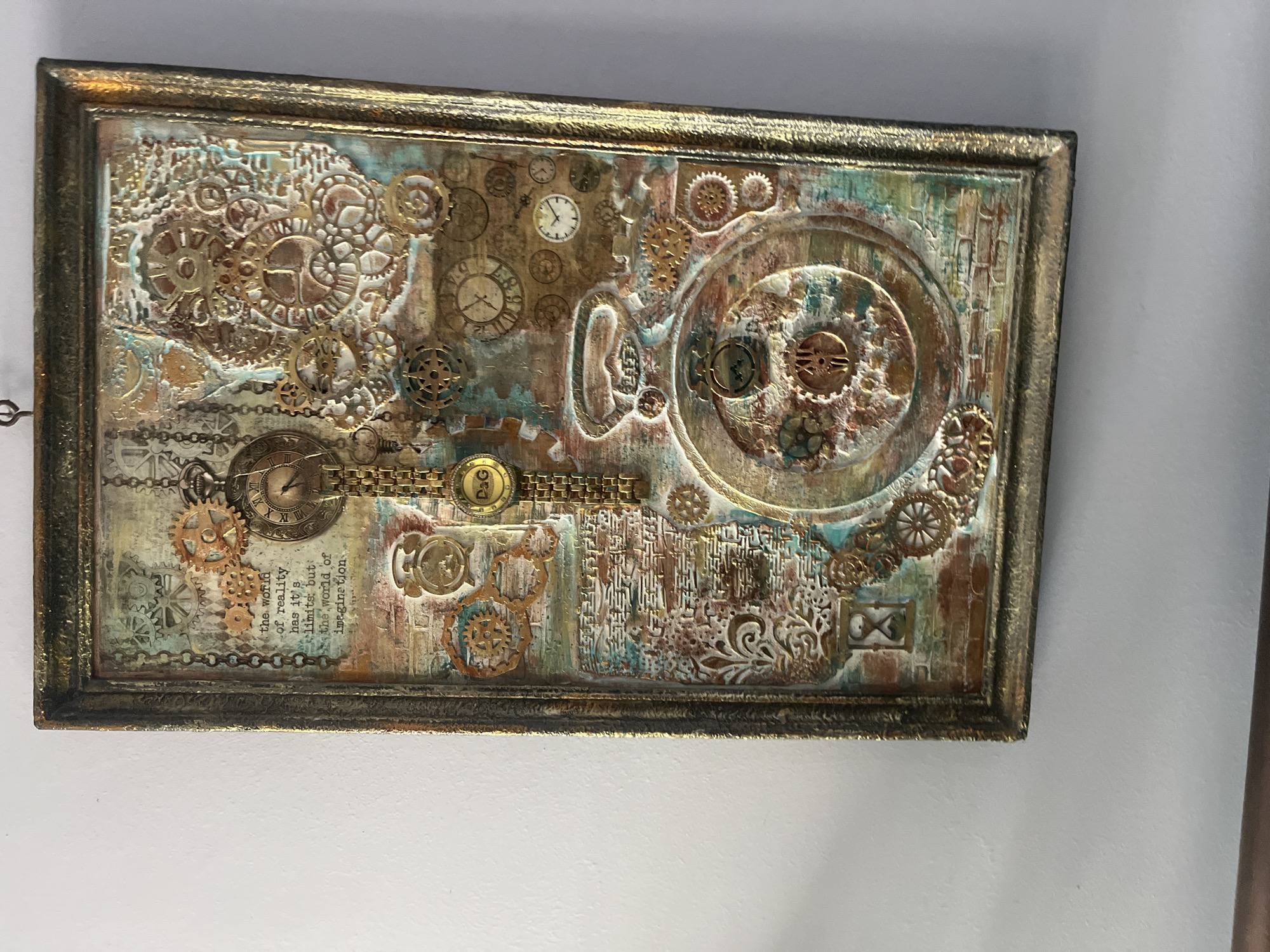 The image shows a framed piece of art hanging on a white wall. The artwork itself is a mixed media collage in a steampunk style. It incorporates various gears, clock faces, chains, and decorative flourishes in tones of gold, bronze, and copper, accented with touches of turquoise and brown. There's also text visible, seemingly fragments of a quote. The frame is metallic with a dark, aged finish. No people are visible. The location in Goce Delcev, Bulgaria is unknown; the image only shows a section of a wall with the artwork.