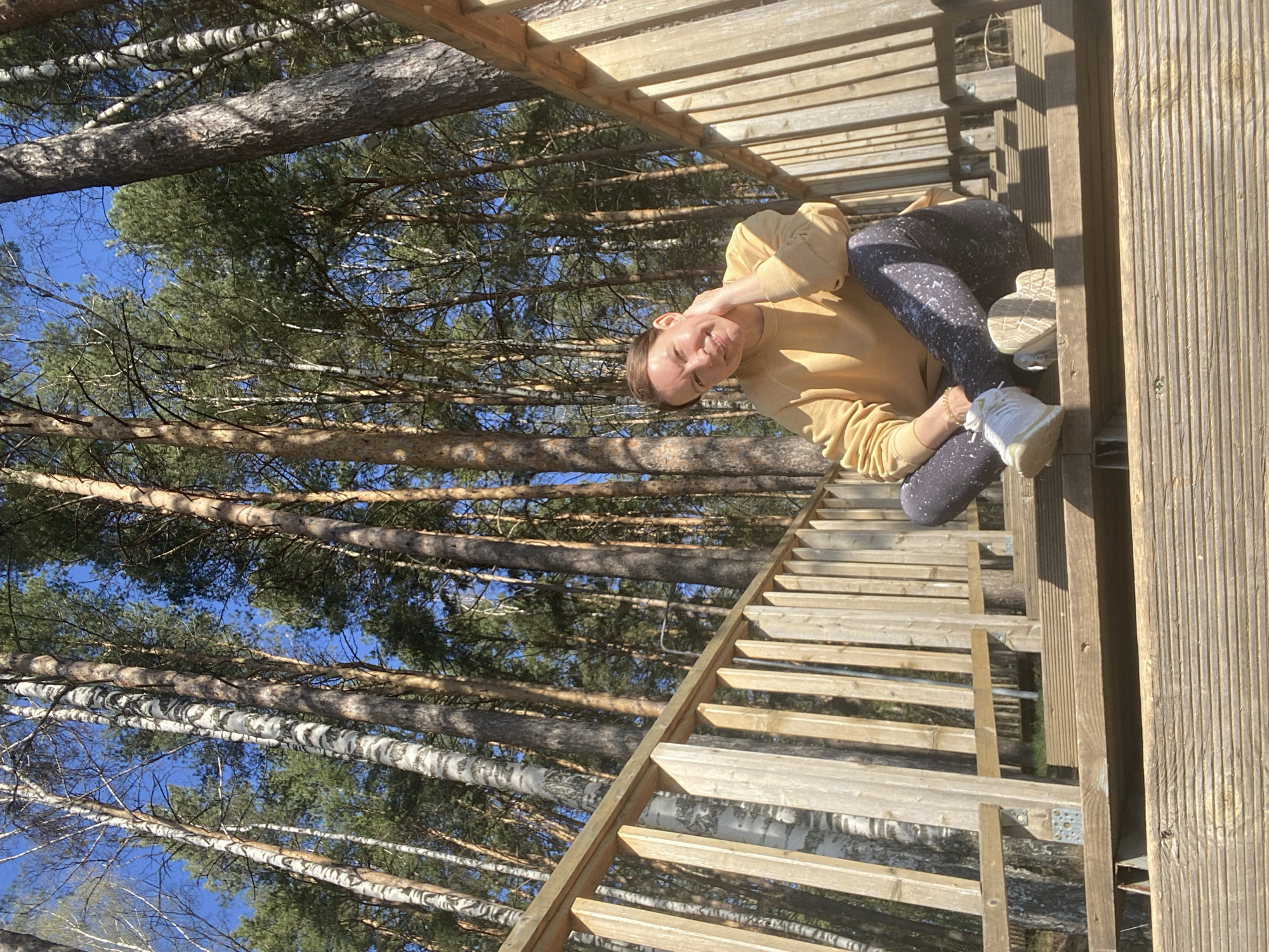 A Caucasian woman is sitting on a wooden structure with a view of tall trees against a blue sky. The structure appears to be a set of stairs or a platform made of light-colored wood. The woman is wearing a tan-colored sweatshirt and patterned leggings. She has light-colored shoes on her feet. The background consists of numerous tall trees, including birch trees with white bark, reaching towards a clear blue sky. The location is Chaikovsky, Russia.