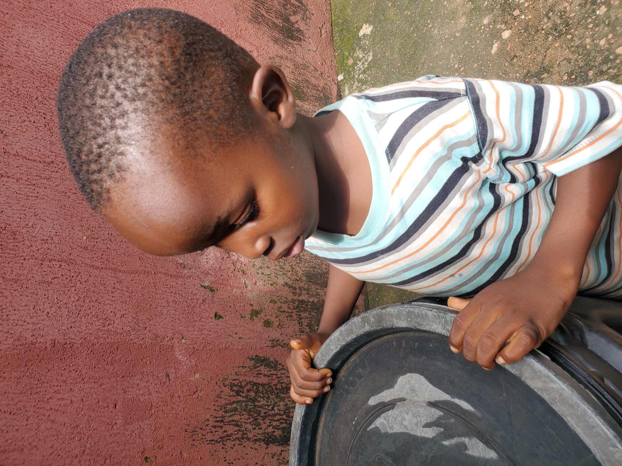 In this medium shot, a young Black child, with closely cropped hair, is shown from a high-angle perspective, looking down with curiosity. The child wears a horizontally striped t-shirt in shades of blue, white, black, and orange. Their right hand rests on the rim of a large, dark-colored plastic container. The container appears to be a basin or tub, possibly containing water, as reflections are visible on its surface.
The background consists of a textured, reddish-brown surface, likely a wall or ground, with patches of green moss and small stones. To the right of the child and the container, a gritty, lighter-colored surface with scattered stones and what appears to be dirt or sand can be seen. The lighting suggests it is daytime, with shadows indicating the sun is likely overhead or at a moderate angle.
The overall scene evokes a sense of everyday life, possibly outdoors in a residential or communal area. The child's focused gaze and gentle hand placement suggest an act of observation or interaction with the container. The setting hints at a warm climate, consistent with the location context of Jos, Nigeria. There is no visible text in the image.