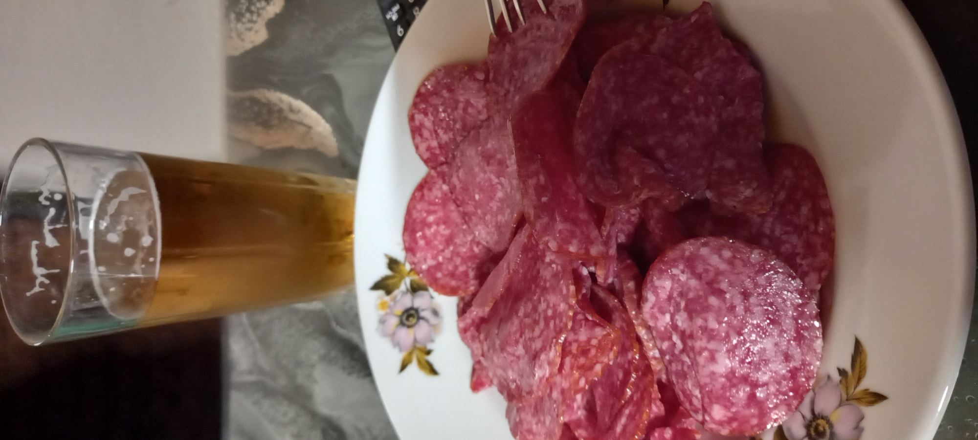 The image depicts a close-up of a plate filled with thinly sliced salami, accompanied by a glass of beer in the background. The salami slices are circular and reddish-pink, with visible white speckles. The plate itself is white and adorned with a floral pattern featuring pink and yellow flowers with green leaves. The glass of beer is tall and transparent, containing a golden-colored liquid with a foamy head. The background is out of focus, showing a dark surface and a patterned tablecloth or surface. There are no people or specific urban landmarks visible that would indicate a location in Iskra, Bulgaria. The setting appears to be indoors, possibly a dining table or a casual setting.
