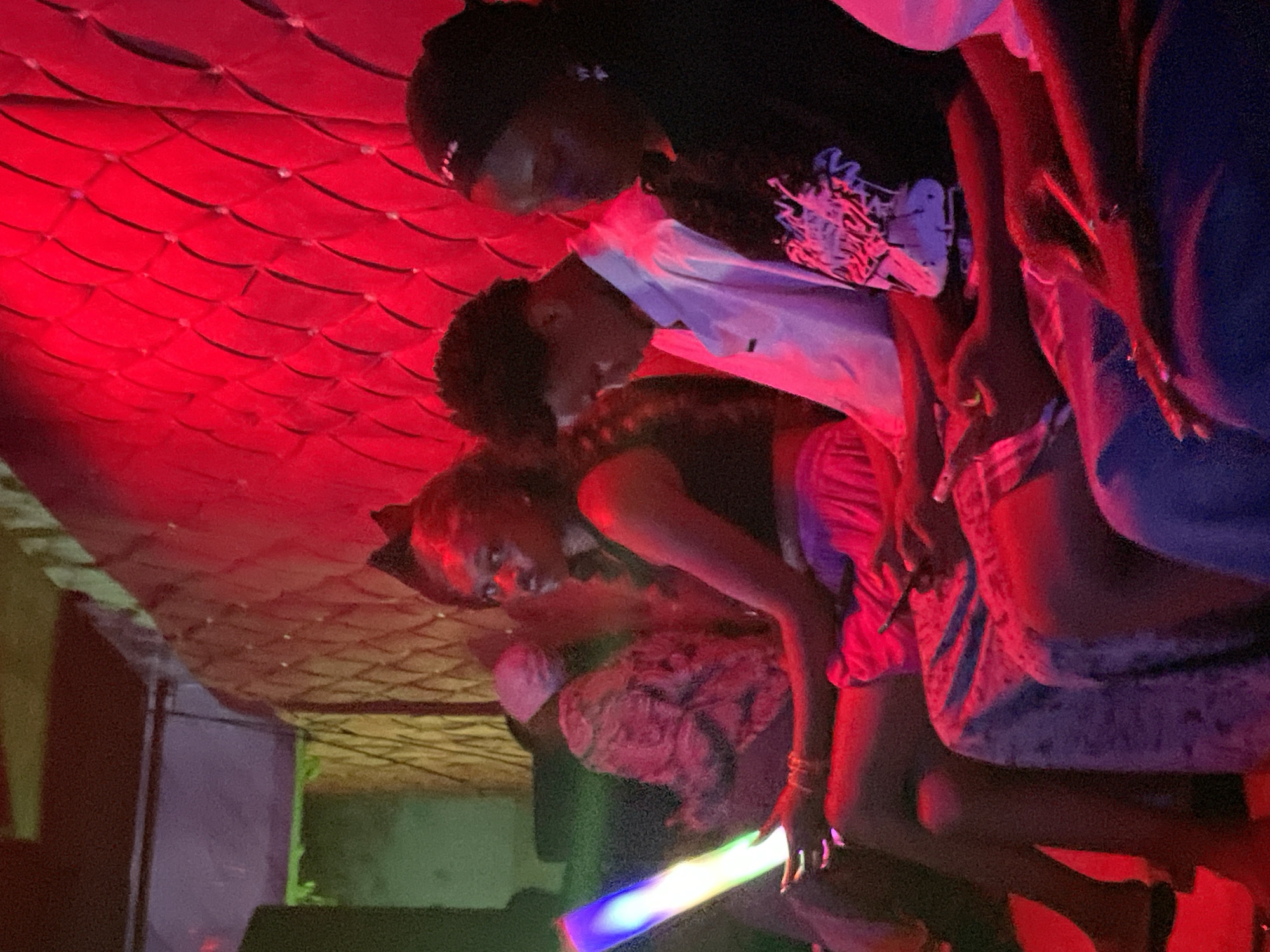 In Durumi, Nigeria, a group of people are seated in what appears to be a dimly lit entertainment venue, possibly a club or lounge. The environment is dominated by vibrant red lighting, which casts a dramatic glow on the surroundings and the individuals. The wall behind the seated individuals features a textured, diamond-tufted pattern, illuminated by the red lights.
In the foreground, a young woman is prominently featured, holding a glowing light stick that displays a spectrum of colors, including blue, green, yellow, and purple. She is wearing a dark top and a light-colored skirt. To her left, another person is partially visible, wearing a patterned top and a pink head covering.
Further to the right, a young man is seated, looking downwards. He is wearing a white collared shirt. Next to him, another young woman is seated, wearing a black t-shirt with a graphic design and a headband. Her gaze is directed away from the camera. The lower portion of the image shows the seated individuals' legs and the surfaces they are sitting on, which appear to be upholstered in a blue fabric. The overall atmosphere suggests a lively and social gathering, possibly at night.