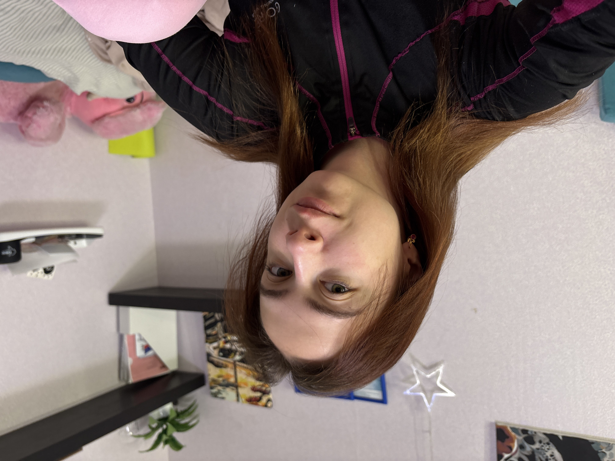 A woman with auburn hair, fair skin, and light eyes is centered in the image. She is wearing a black jacket with magenta trim. The background includes a shelf with books, a small potted plant, and a star-shaped light fixture. A pink plush toy is partially visible. The image appears to have been taken indoors, potentially in a residential space. There is no indication of the location being Прикамский, Russia, or any specific city.