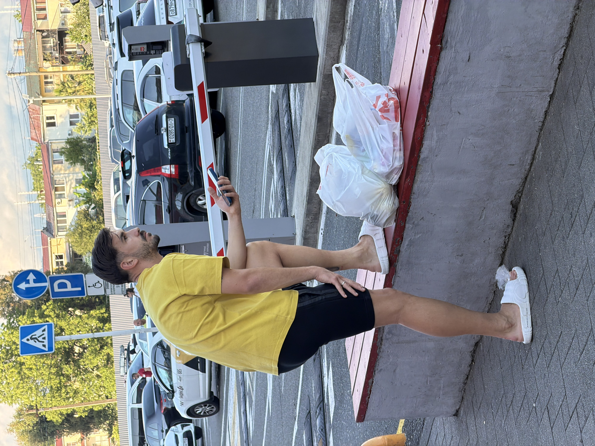 The image shows a man sitting on a red bench next to a parking lot barrier. He is wearing a yellow t-shirt and black shorts, and white slide sandals. The man is holding a smartphone in his hand, and appears to be looking at it. Next to him on the bench are two white plastic shopping bags, one of which contains items. In the background, several cars are parked, and some buildings and trees are visible. There are also several road signs, including a pedestrian crossing sign and a parking sign. The scene appears to be outdoors, likely in a parking lot. The lighting suggests it is daytime, possibly late afternoon or early evening given the warm tones. Visible text includes "0285 01-7" on the license plate of a black SUV and "Яндекс Go" and "885" on a white taxi. The overall atmosphere is casual and relaxed. The setting appears to be Sirius, Russia, as indicated by the provided location context.