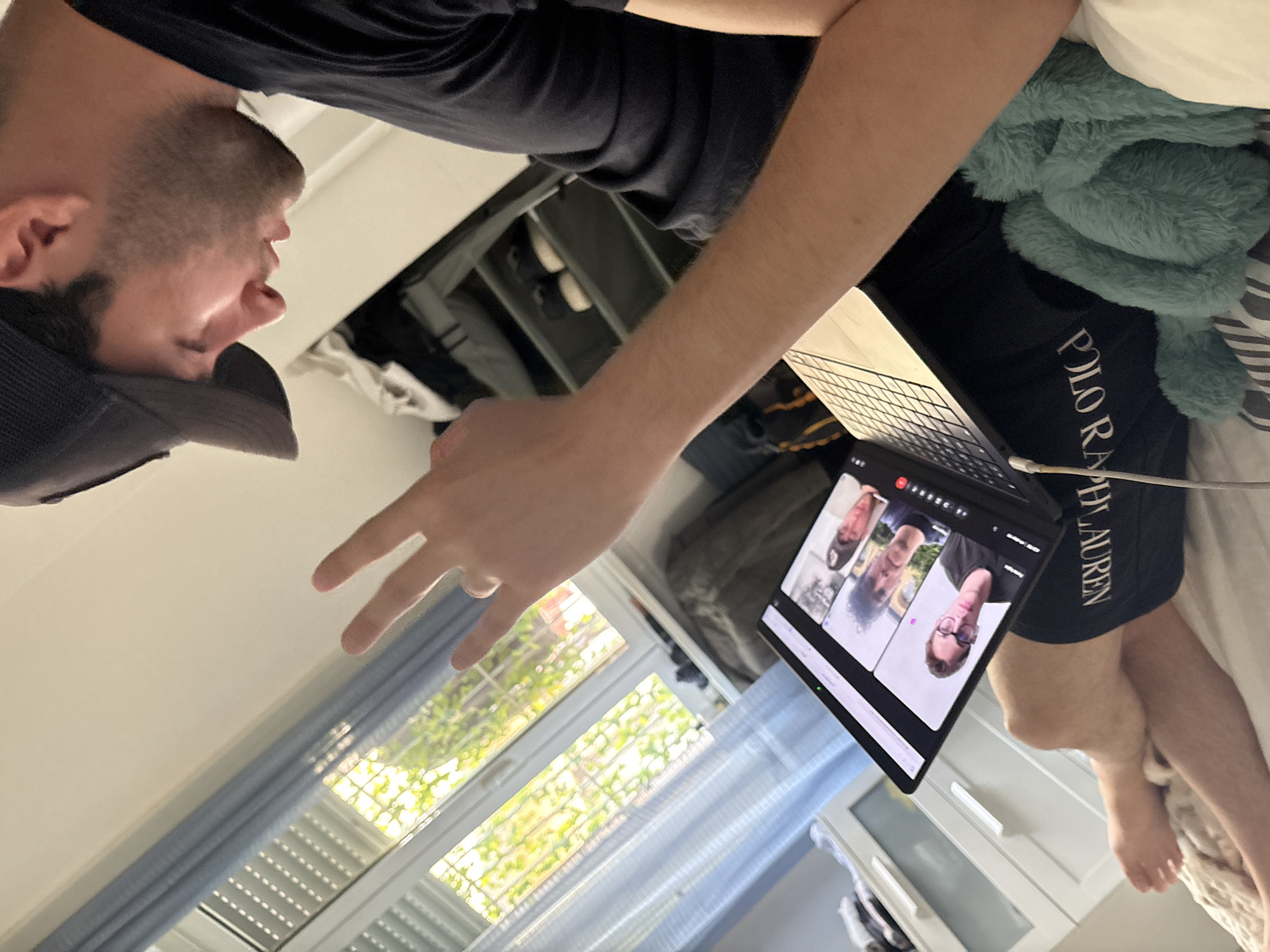 A man is on a video call on a laptop. He is lying on a bed with a stuffed animal next to him. The bed is in a room with a window, and outside the window there are trees and foliage. The location is Milan, Italy.