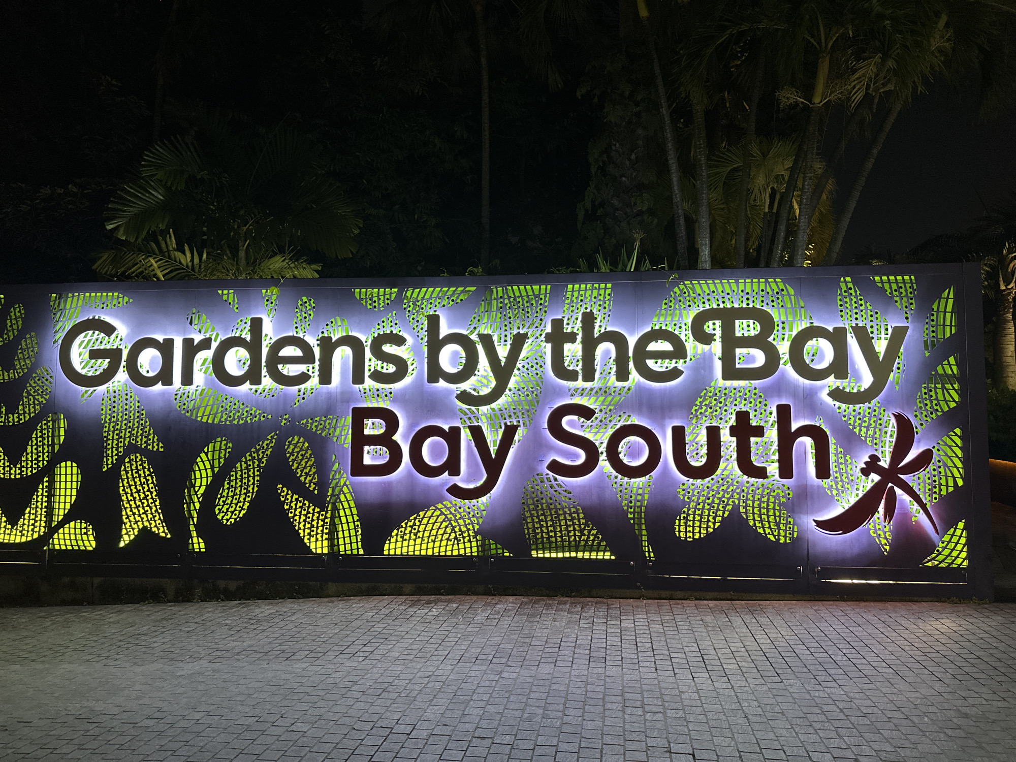 A nighttime, eye-level, full shot captures the illuminated sign for "Gardens by the Bay South" in Singapore. The sign features large, white, backlit letters spelling out "Gardens by the" on the top line, and "Bay South" on the bottom line. The letters are set against a dark panel decorated with a green, leaf-like pattern. To the right of the word "South" is a stylized, dark red dragonfly silhouette. The sign is supported by a black metal frame. The ground in front of the sign is paved with gray cobblestones. In the background, silhouetted palm trees and foliage are visible against the dark night sky. The overall lighting is dim, with the sign being the primary light source.