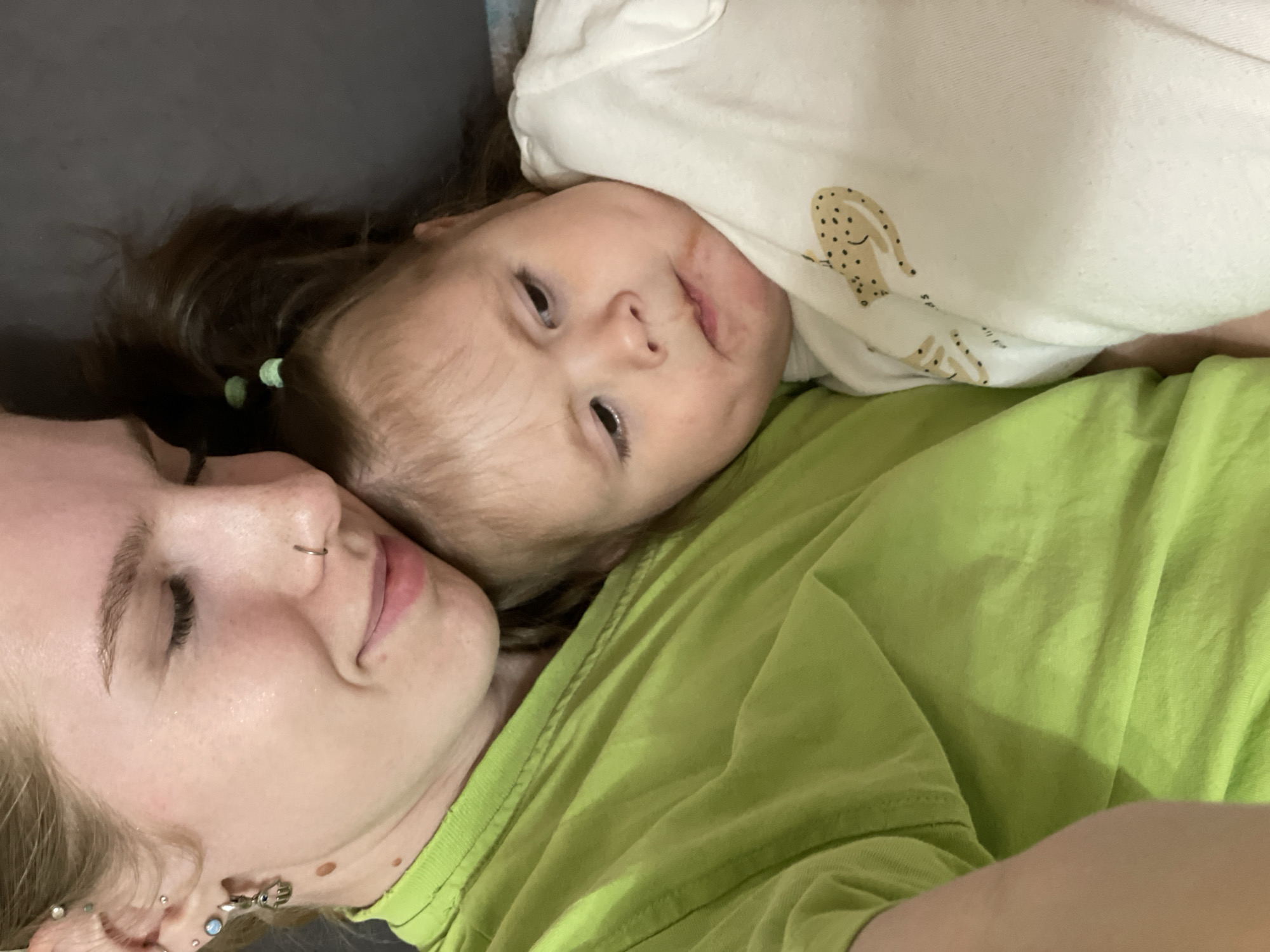 This photograph, taken in Izhevsk, Russia, is initially presented rotated 90 degrees counter-clockwise. When viewed upright, it captures an intimate moment between an adult female and a young child, lying side-by-side.
The adult is positioned on the left side of the frame. She has light-colored hair and appears to be resting, with her eyes closed or looking downwards, revealing a gentle smile. She wears a silver nose ring on her right nostril and several ear piercings, including a distinctive silver earring shaped like a hand. She is dressed in a bright lime green t-shirt.
On the right side of the frame, lying close to the adult, is a young child with dark hair, secured with small green hair ties. The child looks directly towards the viewer with a neutral yet engaged expression, displaying light eyes. Faint reddish marks are visible on the child's cheek and chin area. The child wears a white top adorned with a light brown, spotted animal design, possibly a bunny or deer, and partially visible text that appears to read "SPRI...".
The setting appears to be indoors, likely a bedroom or living room, with both individuals resting on a dark grey or black surface, possibly a bed or sofa. The soft, ambient lighting suggests it is daytime indoors. The overall mood conveyed is one of tenderness, closeness, and relaxation between the two individuals. No other specific location cues or weather details are discernible from the image.
