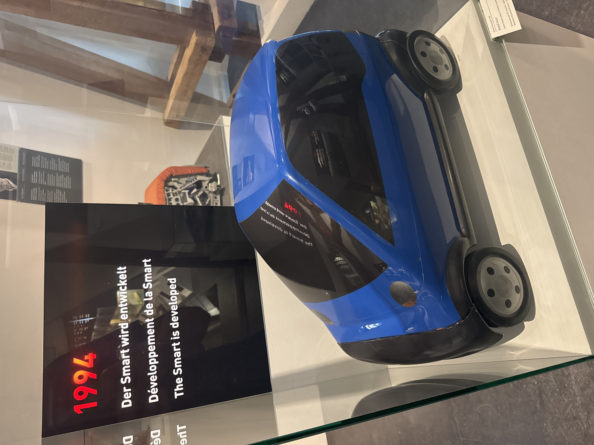 This media file depicts a blue model of a Smart car displayed in a museum exhibit. The car is positioned on its side, showcasing its sleek design and black tinted windows. In the background, wooden beams are visible, suggesting an older architectural setting.
A prominent display panel to the left of the car features text in three languages: German, French, and English, all stating "Der Smart wird entwickelt," "Développement de la Smart," and "The Smart is developed," respectively. Above these phrases, the number "1994" is displayed in large red font, indicating the year associated with the Smart car's development. Further text on the display panel, partially obscured, appears to provide historical context related to the car's model and its manufacturing origins in Grenchen, Switzerland, with a reference to the "Berna Historical Museum."
The scene is well-lit, likely within an indoor museum environment. There are no people visible in the immediate frame, focusing the attention entirely on the exhibit. The overall impression is one of historical preservation and presentation of an iconic automotive design.