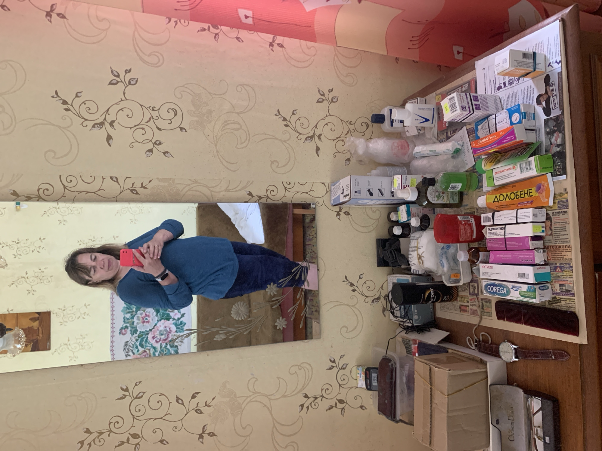 A woman stands in front of a mirror, taking a selfie with a red smartphone. She is wearing a blue long-sleeved shirt and dark blue pants. The mirror reflects her upper body and lower body. The background of the image shows a wall with floral wallpaper and a curtain. On a table in front of the mirror, there are various items, including boxes, bottles, a watch, and a comb. The woman appears to be in a room, likely a bedroom or living area. Based on the context of "Відзаўскі сельскі Савет, Belarus", the location is a rural area in Belarus.