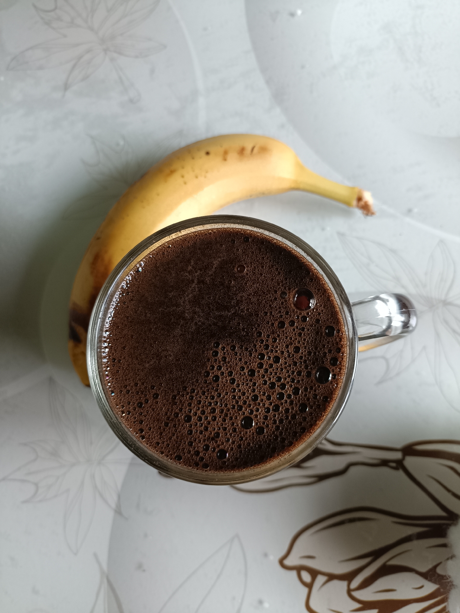A top-down, close-up shot features a clear glass mug filled with dark, frothy coffee, positioned on a light-colored surface adorned with a subtle floral pattern. A single, slightly curved, yellow banana with a few brown spots rests behind the mug. The scene is captured indoors, likely on a table, with soft, diffused lighting. The background is a blur of light grays and whites, with faint floral motifs visible on the surface. No people are present in the image. The location of Bataysk, Russia, is not discernible from the visual information provided.