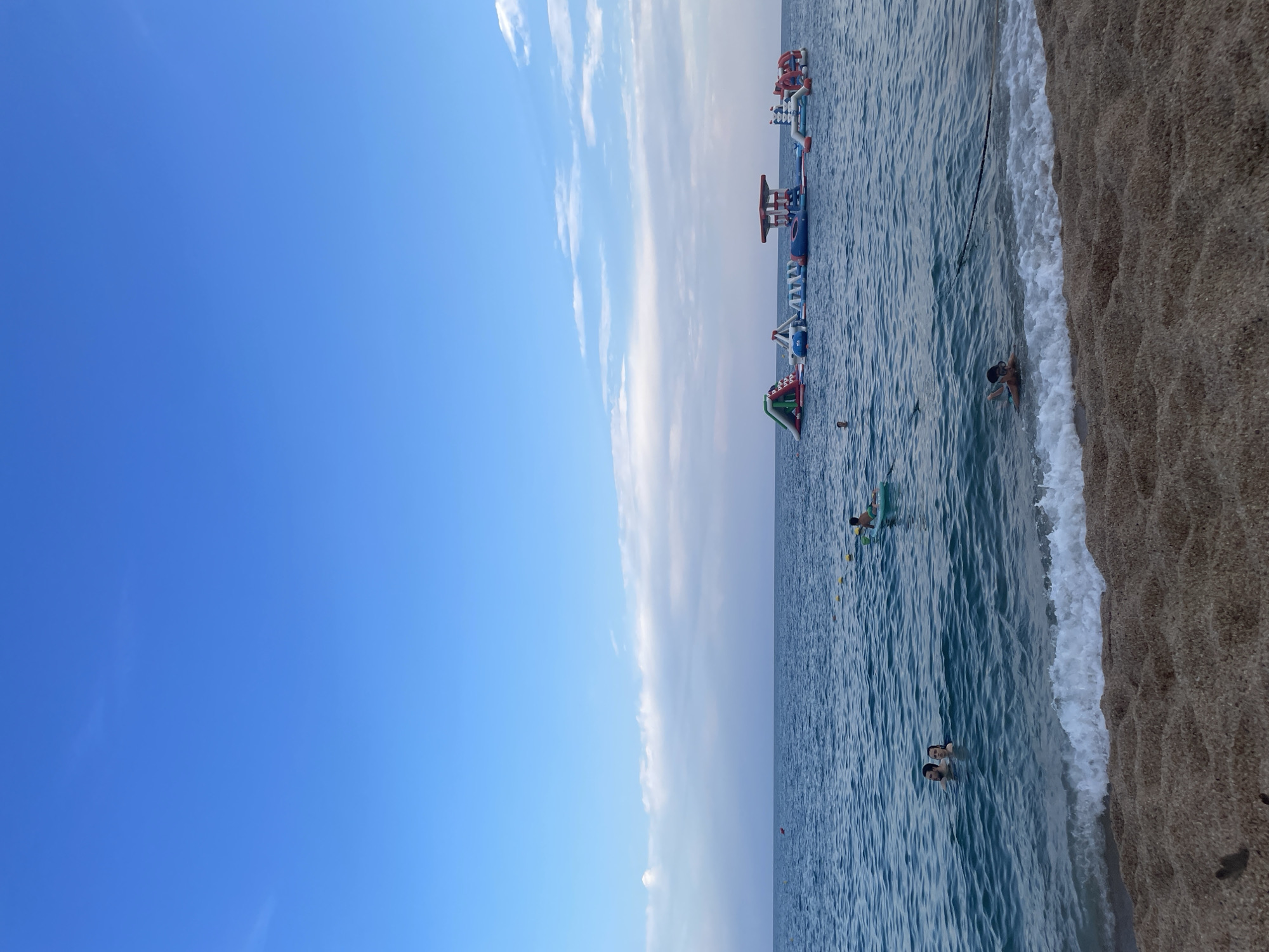 The image depicts a sunny day at the beach in Canet de Mar, Spain. The foreground shows a sandy beach with gentle waves lapping at the shore. In the middle ground, the blue ocean is populated with several people swimming and playing. Two individuals are close to the shore, their heads visible above the water. Further out, a young person is lying on a float. In the background, a large inflatable water park structure with slides and platforms is anchored in the sea. The sky above is a clear blue with scattered white clouds.