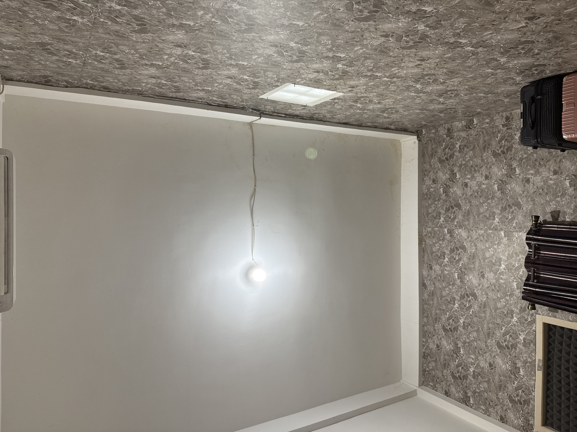 This image depicts an interior shot of a room, likely a bedroom or hotel room, with a view angled towards the ceiling and two adjacent walls. The ceiling is white and appears to be made of a smooth material, possibly gypsum board, with a single, bare light bulb illuminating the space. A thin electrical wire runs horizontally across the ceiling, attached at intervals. An air conditioning unit is visible at the top edge of the frame, partially obscured.
One wall is covered in a patterned wallpaper with a gray, marbled design. A small, square window with a translucent covering is set into this wall. The other wall, visible on the left, is also covered in the same marbled wallpaper.
In the lower portion of the image, on the right side, a stack of two suitcases is visible: a black hard-shell suitcase on top of a pink one. On the left side, a window frame with a textured acoustic panel behind it is partially visible, and next to it, a set of dark curtains with vertical stripes.
The overall impression is that of a functional, perhaps budget-friendly, lodging space. The image does not provide specific visual cues to pinpoint its exact location within Bandar Lampung, Indonesia, beyond being an indoor setting.