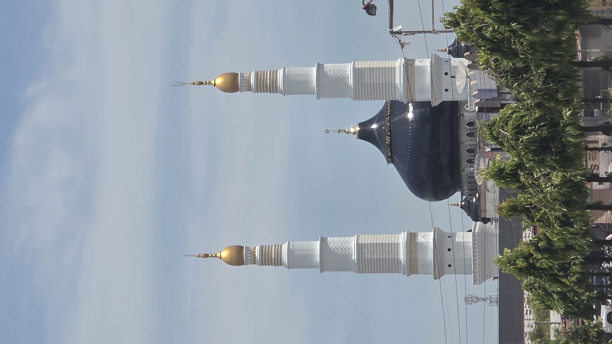 The image shows the Baiturrahman Grand Mosque in Pantegajah, Indonesia. Two white minarets with golden spires flank a large, black dome with a golden spire. Green trees are visible on the right side of the image, obscuring the lower portion of the mosque. The sky is light blue with no clouds. Utility poles and wires are also visible.