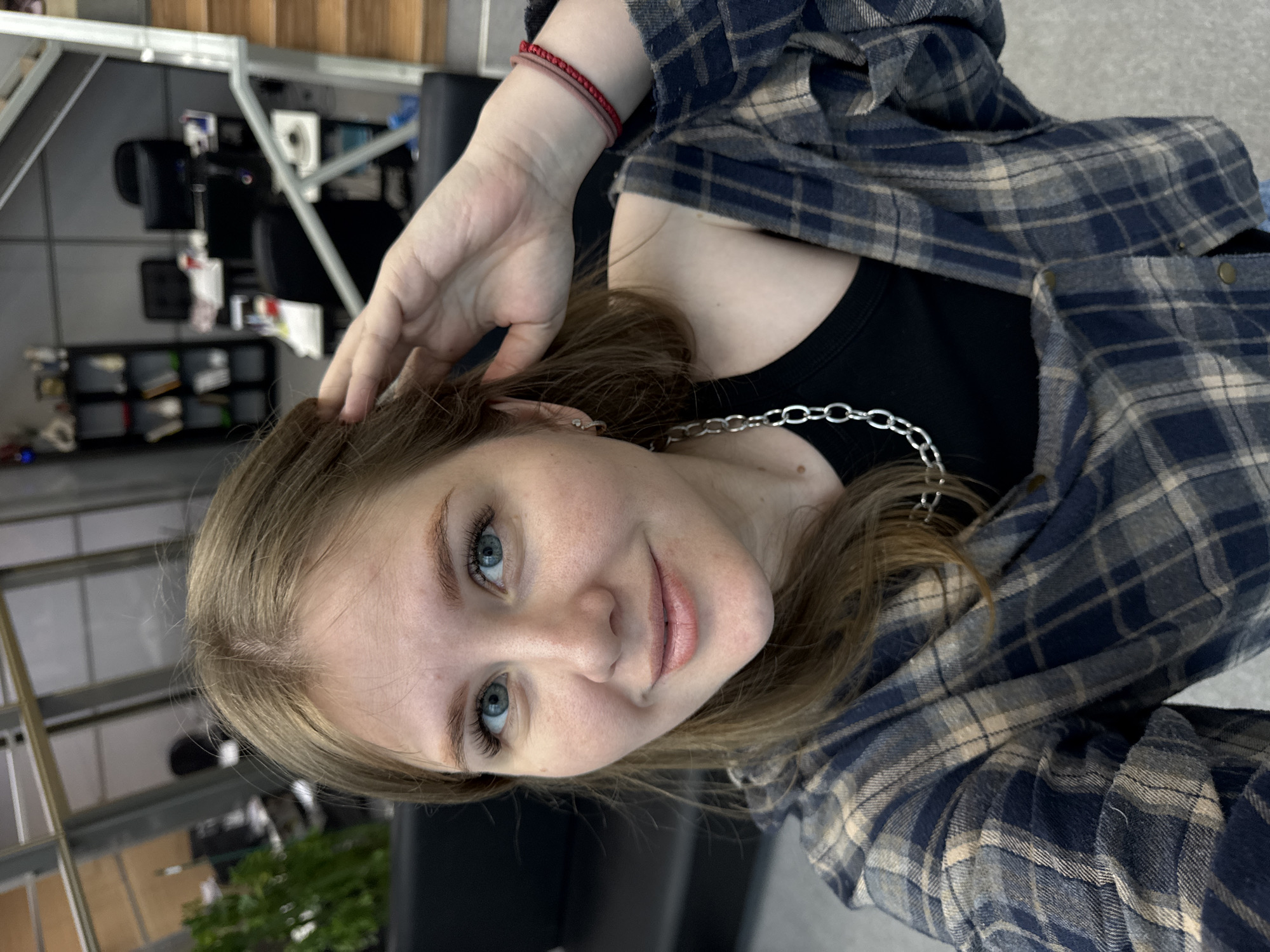 A young woman, likely in her late teens or early twenties, is depicted in a selfie, looking directly at the camera with a soft smile. She has light brown hair, fair skin, and striking blue eyes with prominent eyelashes. Her expression is calm and friendly.
She is dressed in a black top, possibly a tank top, featuring a silver chain necklace. Over this, she wears an open blue and beige plaid flannel shirt. Two thin red string bracelets adorn her right wrist. Her right hand is raised, with fingers gently touching her hair near her head, a common selfie pose.
The background appears to be an indoor setting, potentially a salon, office, or public space, characterized by dark furniture, possibly chairs, and shelves holding various items. Metallic structures are also visible in the background, along with a small green plant on the left side of the frame. The lighting suggests an indoor environment, likely artificial. The image is oriented 90 degrees counter-clockwise. No text is visible in the media. The contextual location is Perm, Russia.