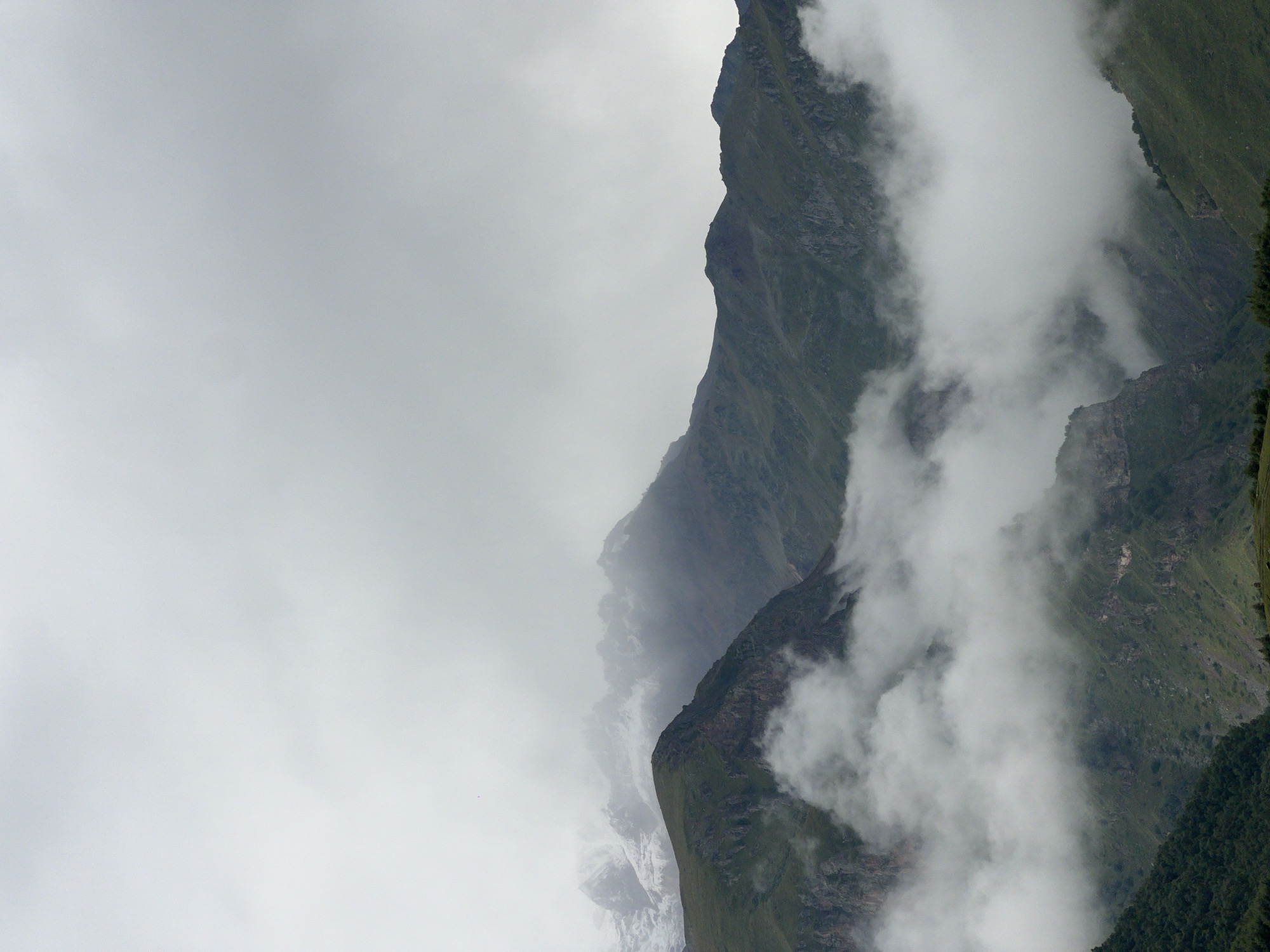 A wide shot captures a moody, overcast day in Gergeti, Georgia, with a focus on layered mountain ranges partially obscured by thick, white fog. In the foreground, a dark, rocky mountain peak covered in green grass dominates the left side. Behind it, a second, slightly taller mountain range, also green and rocky, is visible, with snow-capped peaks further in the distance peaking through the clouds on the far left. The middle ground features a dramatic flow of fog swirling around the slopes, creating depth and a sense of mystery. The background is a hazy expanse of gray sky blending into the distant mountains, with hints of darker, more rugged terrain visible through breaks in the mist. The overall impression is one of dramatic natural beauty and a sense of being immersed in the elements, with no people or man-made structures evident.