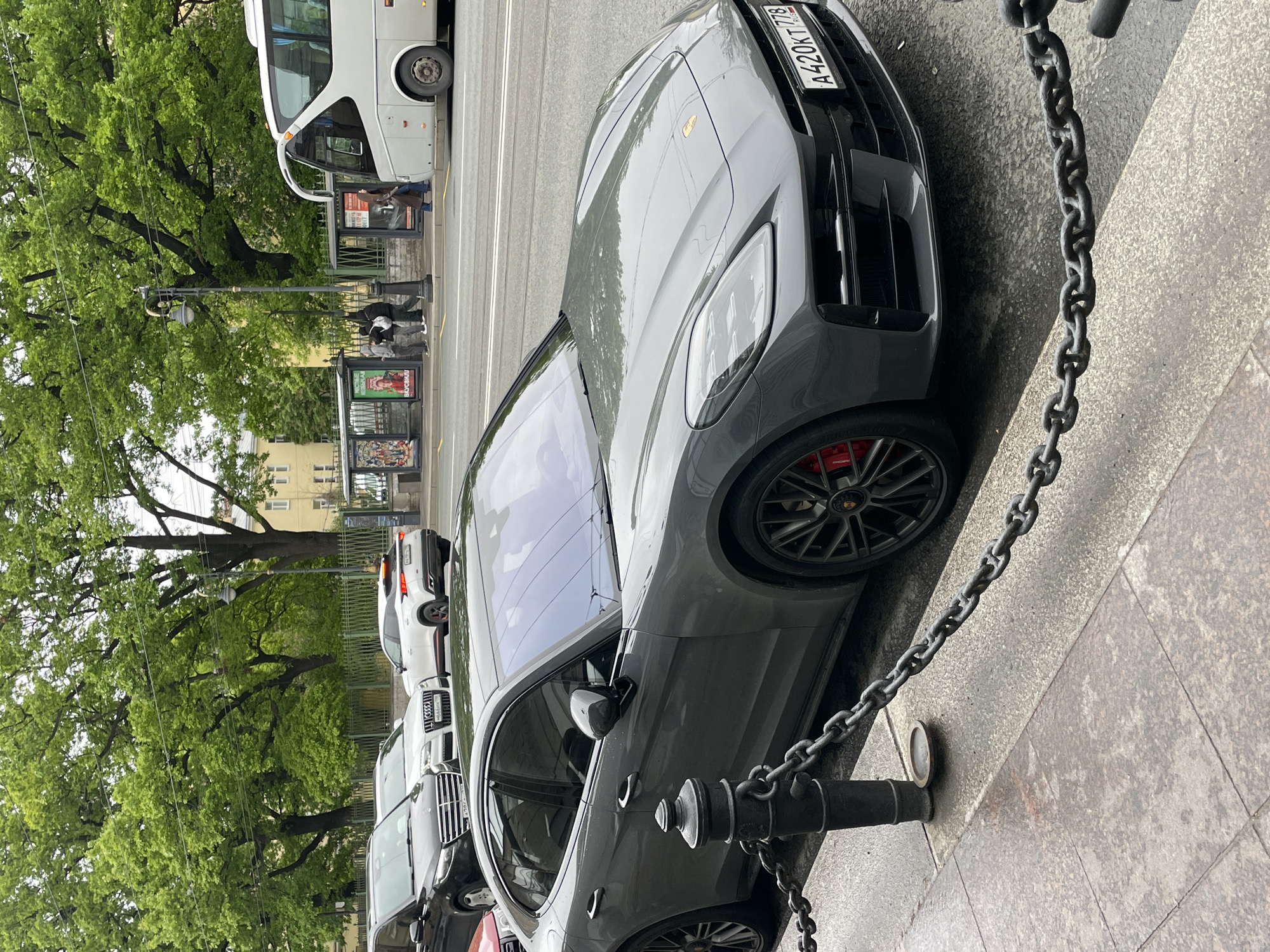 Here is a detailed description of the image:
The image captures a street scene in Saint Petersburg, Russia, showcasing a gray Porsche parked along the sidewalk adjacent to a chain barrier. The license plate on the Porsche reads "A420KT778".
Along the street, a passenger bus can be seen, and behind the Porsche, other parked cars are visible. A bus stop with visible advertisements stands further down the street. In the background, lush green trees line the sidewalk, providing a natural backdrop to the urban environment. A light-colored building is discernible behind the trees. Several pedestrians are present near the bus stop.