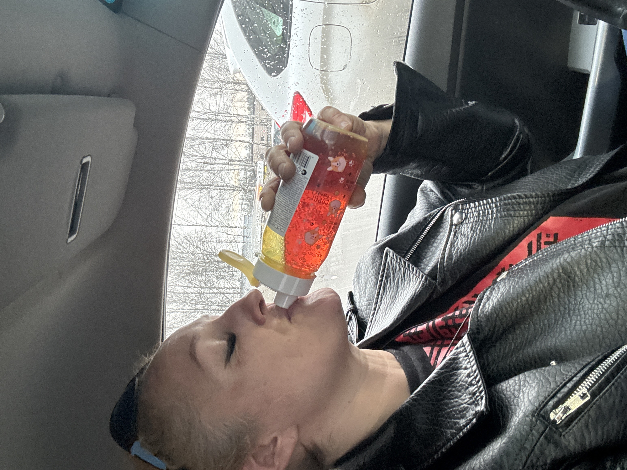 A fair-skinned woman is inside a car, drinking from a transparent, plastic bottle containing a red liquid. The bottle has cartoon corgi images on it. The woman is wearing a black leather jacket over a red and black top. Raindrops are visible on the car's windshield. Through the windshield, trees with bare branches are visible. The location is Izhevsk, Russia.