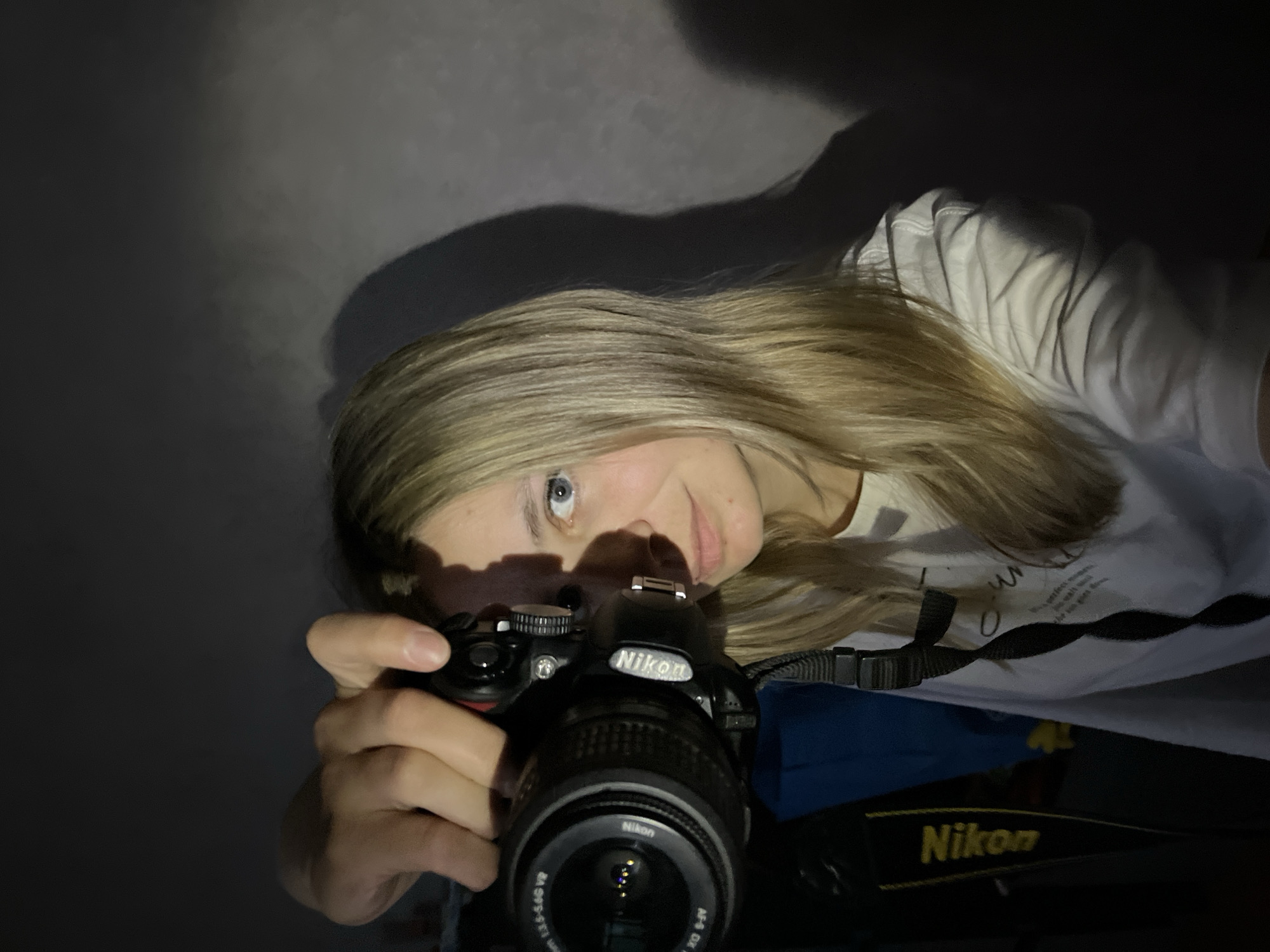 A woman with blonde hair holds a Nikon DSLR camera. She is looking at the camera, and her hand is on the shutter. The camera is focused, and the background is dark. The text on her white t-shirt is partially visible: “The perfect moments …”. Another portion of Nikon camera hardware is on the bottom right.
I cannot confirm the location based on the given information.