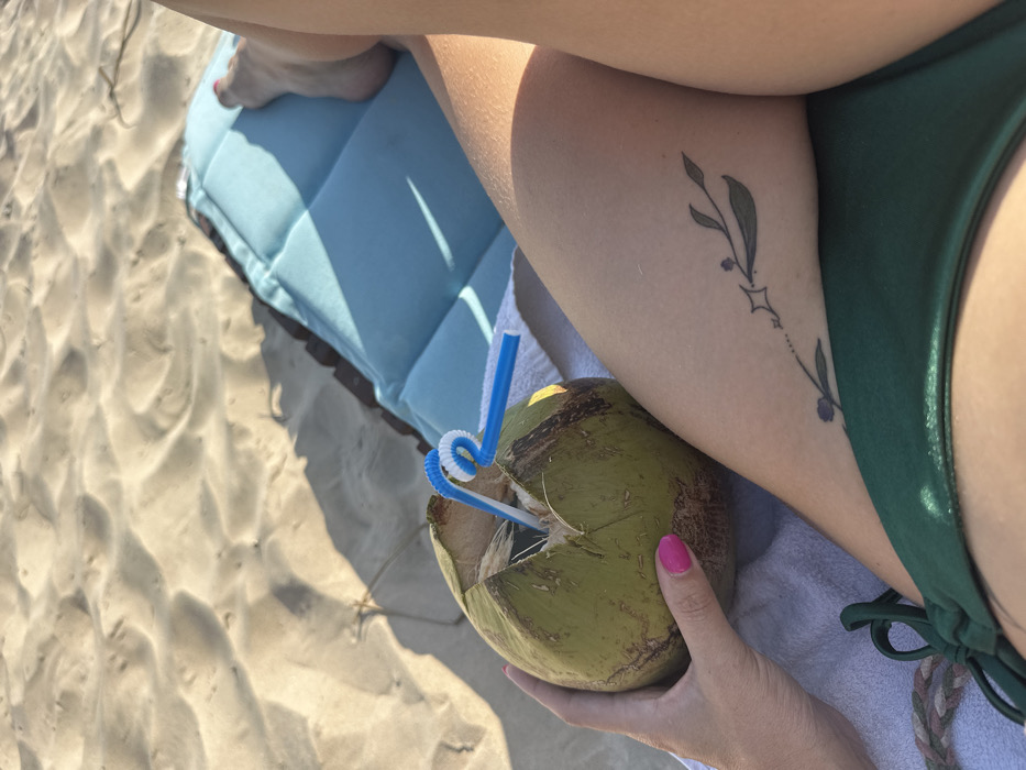 The scene depicts a person relaxing on a sandy beach in Thành phố Phan Thiết, Vietnam, under bright daylight. The upper portion of the image shows light-colored sand with ripple patterns and distinct shadows. In the foreground, a person's body is partially visible, primarily focusing on their legs, left hand, and midsection. The left hand, with pink-painted fingernails, holds a green coconut, which has been opened at the top and contains two plastic straws: one blue and white striped, the other plain blue. A white towel or fabric rests on the person's lap beneath the coconut. The person's right upper thigh features a small tattoo depicting a stylized vine with leaves, dots, and a four-pointed star. A dark green bikini bottom with visible ties covers the lower midsection. A light blue quilted beach mat or cushion is positioned beneath the person's legs, resting on the sand. The person's right foot, with red-painted toenails, is visible near the upper right corner of the mat.