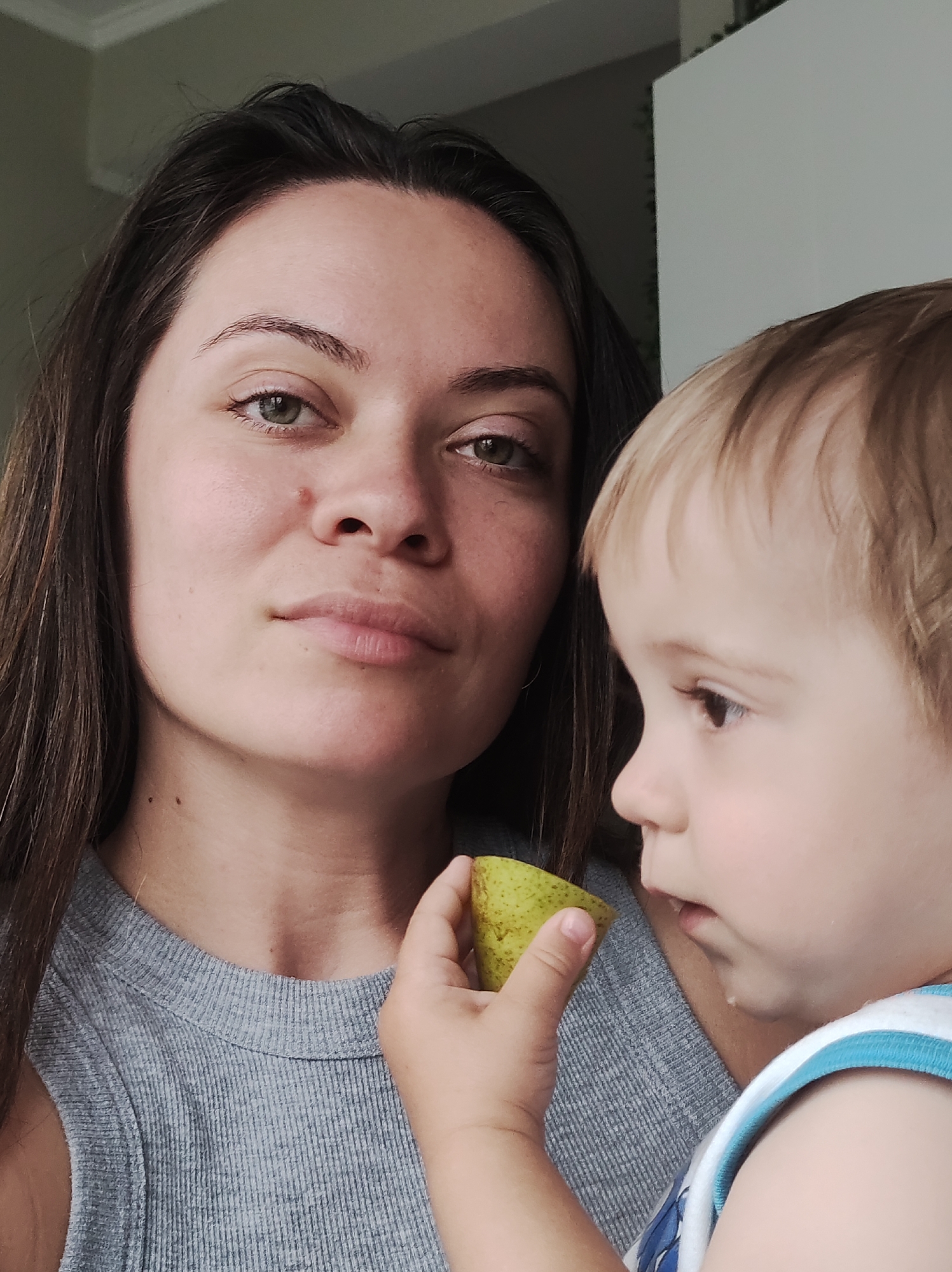This media file captures an intimate, close-up indoor portrait of an adult woman and a young child, likely a mother and her child, in Chaikovsky, Russia.
The woman occupies the left and central portion of the frame, looking directly at the viewer with a neutral, slightly serious, or contemplative expression. She has long, dark brown hair and striking green or hazel eyes. Her skin appears clear, and she wears a simple grey, ribbed sleeveless top.
The young child is positioned on the right, partially visible from the side profile, with light blonde or light brown hair. The child's right hand holds a light green pear, which shows signs of being partially eaten. The child is looking towards the right, away from the camera, seemingly focused on the fruit, with a small trace of liquid or juice on their chin. The child wears a white sleeveless top with blue stripes.
The background is softly blurred, suggesting an indoor setting with subtle ambient light, possibly from a window, indicating daytime. The colors are natural and subdued, highlighting the subjects. No specific location cues beyond the general indoor setting are visible, nor is any text present in the image. The photograph captures a quiet, everyday moment between the two individuals.