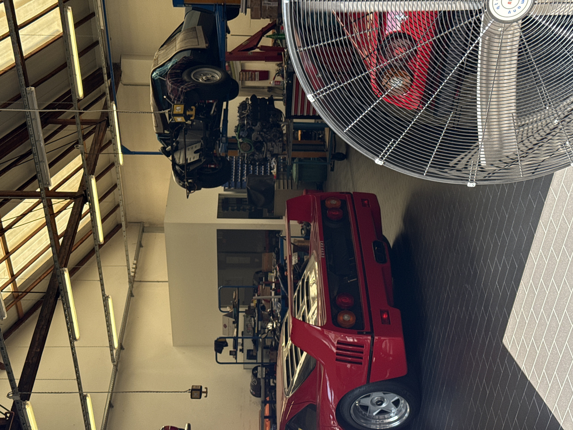 The image shows the interior of a car repair workshop. In the foreground on the left is the rear of a red Ferrari F40. In the middle ground, another car, possibly a black Ferrari, is suspended on a car lift. An engine is visible on a trolley below the lifted car. The workshop has a high ceiling with exposed beams and fluorescent lighting. Shelving units with tools and car parts are visible on the walls. A large industrial fan is positioned in the right foreground, partially obscuring the scene. The floor is tiled. There are no people visible in the image. The location is identified as a car workshop in Offenbach am Main, Germany.