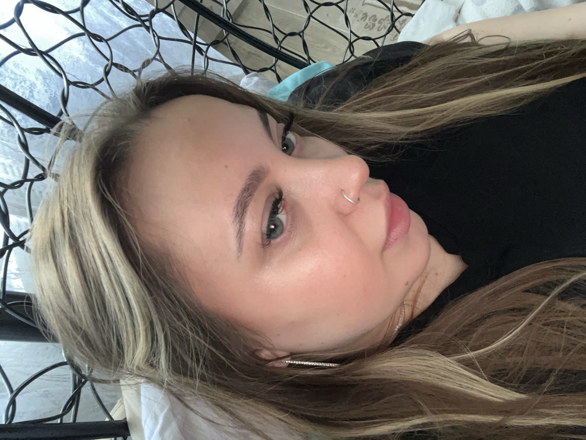 A person with light skin, light brown hair with blonde highlights, and light-colored eyes is lying down, facing the camera. They are wearing a black shirt and have a silver hoop earring in their ear and a silver nose ring. The background consists of a metal frame structure, a white pillow, and some light-colored fabric. The location is not discernible from the image.