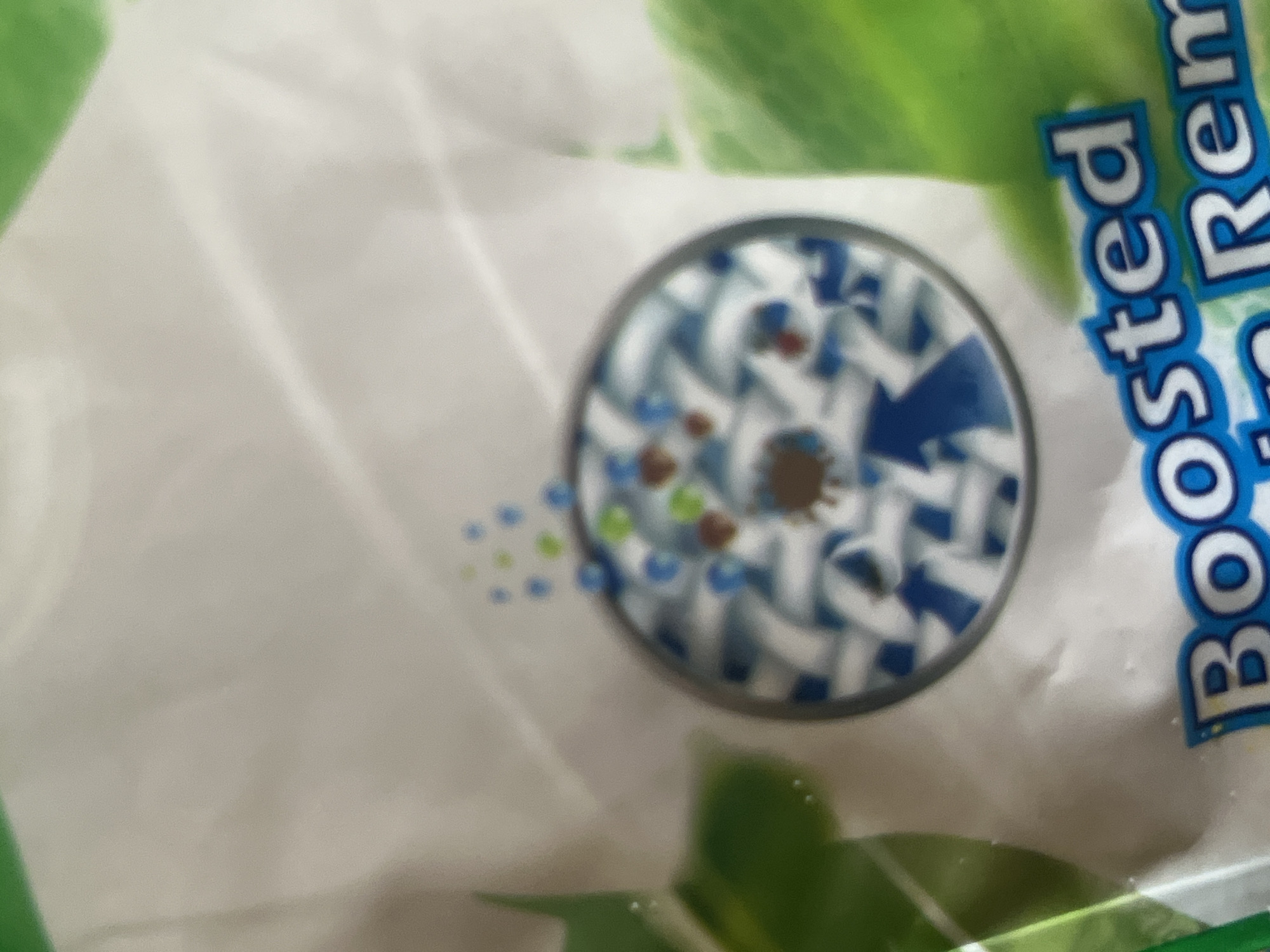 The image shows a close-up of a white t-shirt with a circular graphic depicting a stain being removed from fabric. The graphic shows a close-up of woven fabric with blue and white strands, and some small brown, green, and blue dots representing stains. An arrow indicates the removal of a prominent brown stain in the center of the circle. Below the graphic, text is partially visible, reading "Boosted" and "n Rem". Green leaf-like shapes are present on the right side of the image, suggesting a connection to nature or a product with natural ingredients. The image appears to be a product advertisement or packaging. There are no people visible in the image. The location of Jauro Yinu, Nigeria, is not depicted or suggested by the image content.