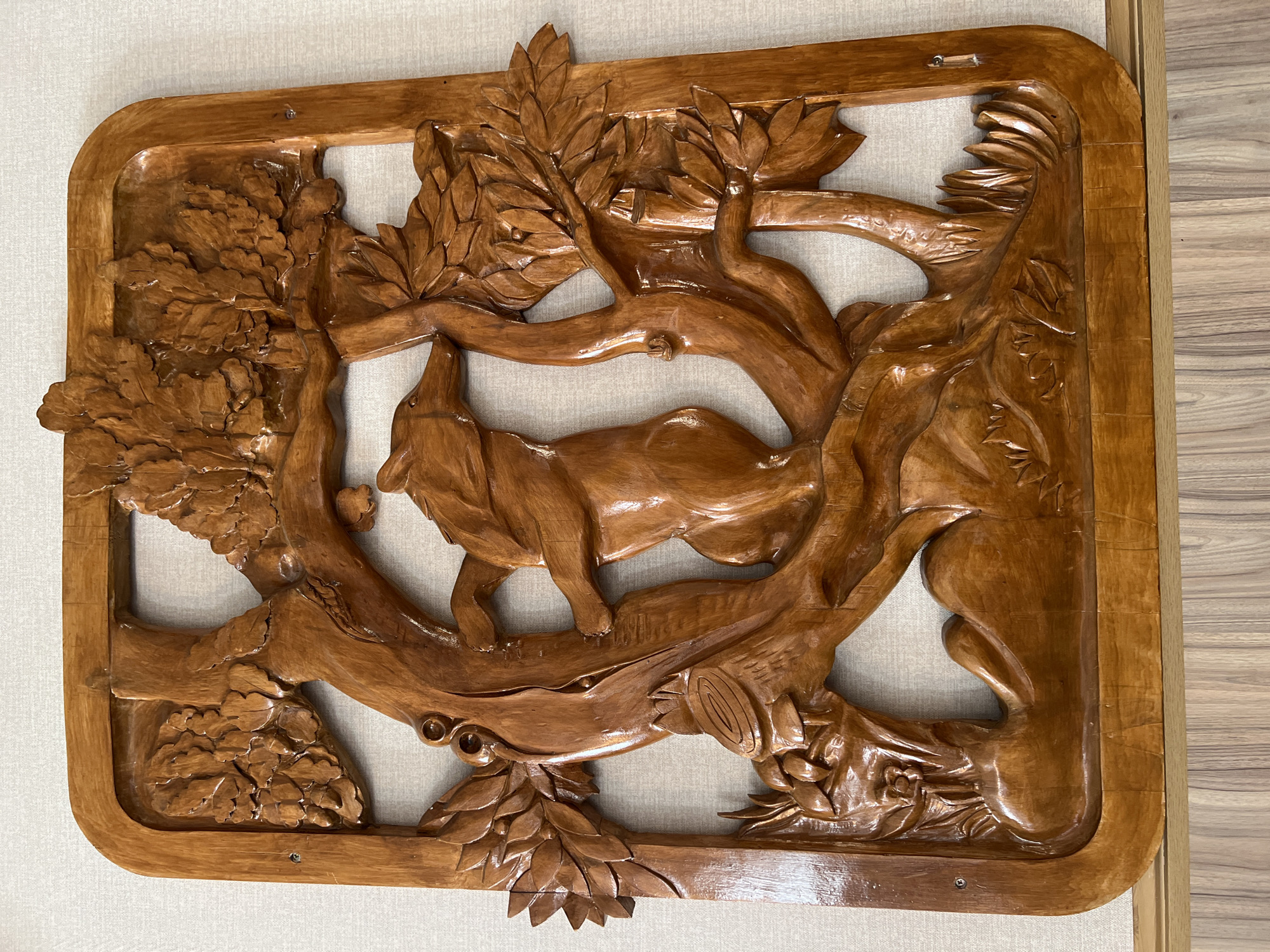 This media file displays a polished, reddish-brown wooden bas-relief carving, likely mounted on a wall. The central subject is a dynamically posed, wolf-like animal, possibly a fox or dog, with canine features and a bushy tail. It appears to be navigating or resting amidst intricately carved trees, branches, and leaves that form a naturalistic setting within the artwork. The animal's head is raised, suggesting it might be howling, sniffing, or looking upwards.
The carving itself is framed by a solid wooden border, with visible screws in the corners, indicating its attachment to a lighter, possibly off-white or beige, textured background material, which could be fabric or a wall. The glossy finish of the wood reflects light, enhancing the depth and detail of the carving, particularly the textures of the leaves and the animal's form. The artwork is set in Sanatoriya Yumatovo Imeni 15-Letiya Bassr, Russia. There is no visible text in the image.