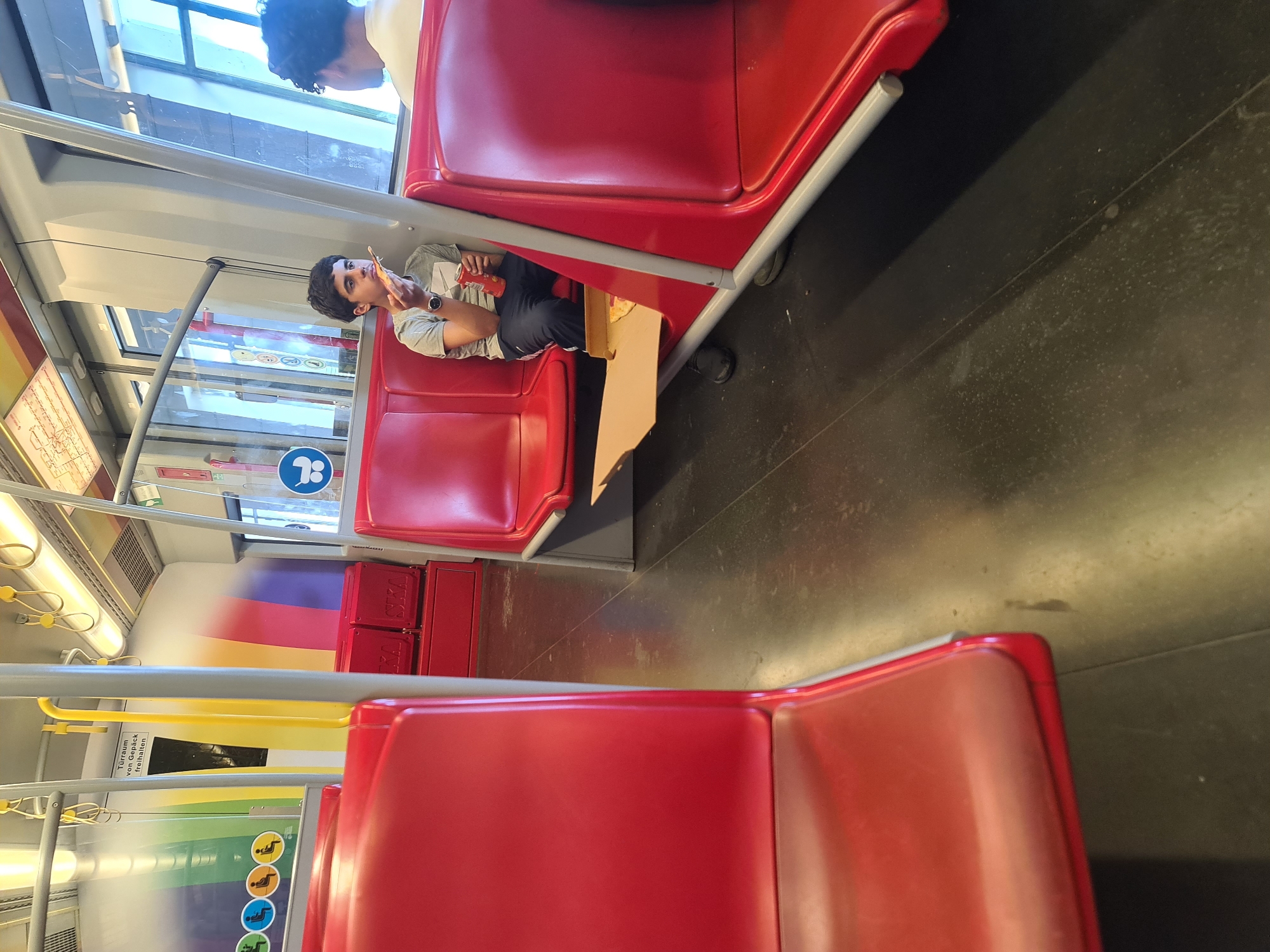 The image depicts the interior of a Vienna U-Bahn train, as evidenced by the distinctive red seating, the overhead handles, and the signage in German ("Türmen von Gepäck freihalten"). In the foreground, two rows of red seats are visible. On the right side of the frame, a young male with dark curly hair is seated, wearing a grey t-shirt and dark pants. He is holding a slice of pizza and a red can. In front of him, a pizza box with one slice of pizza remaining is placed on a lower ledge. To his right, another person is partially visible, facing away from the camera. The floor of the train is dark and appears to be made of a durable material. The walls of the train car feature colorful stripes and informational posters, including one with a rainbow motif and symbols indicating passenger information and seating. The overall impression is a typical scene inside public transportation in Vienna.