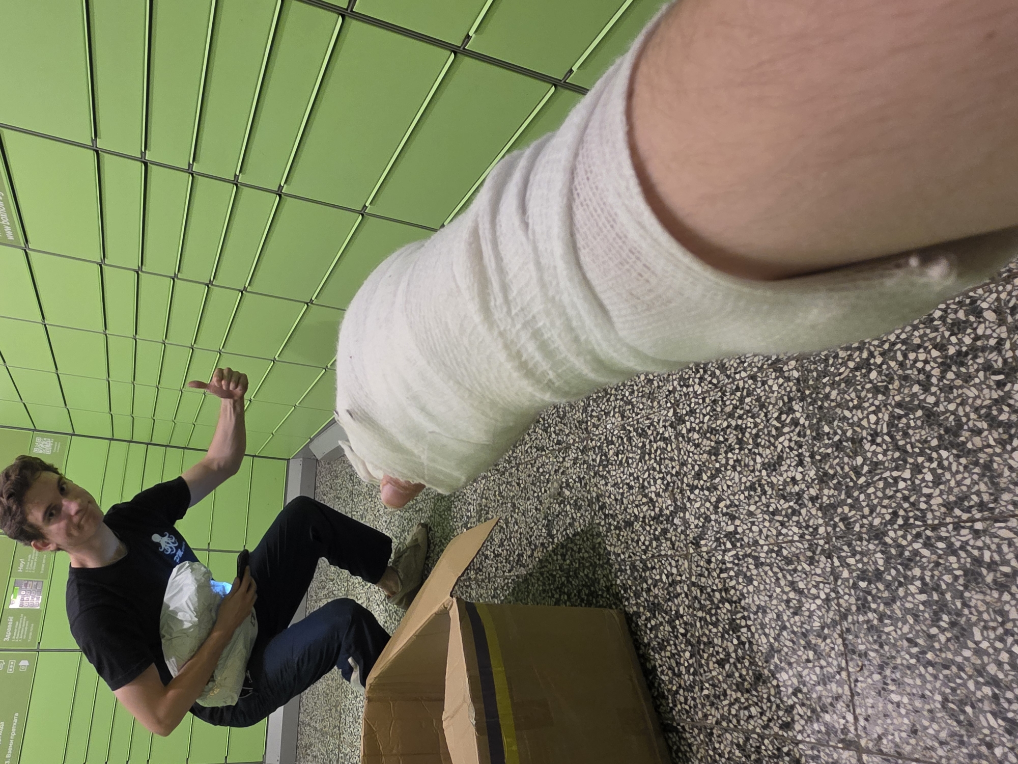 A young man with a cast on his left arm is crouching next to an open cardboard box. He is holding a white package and giving a thumbs-up gesture. The background consists of a wall of green lockers, typical of automated parcel lockers. The floor is tiled with a speckled pattern. The scene is indoors, likely at a parcel locker station. The presence of Bulgarian text on the lockers, specifically "www.boxnow.bg," indicates the location is in Bulgaria, and BoxNow is a common parcel locker service there. The specific image does not provide enough detail to pinpoint a particular street or landmark within Sofia, Bulgaria.