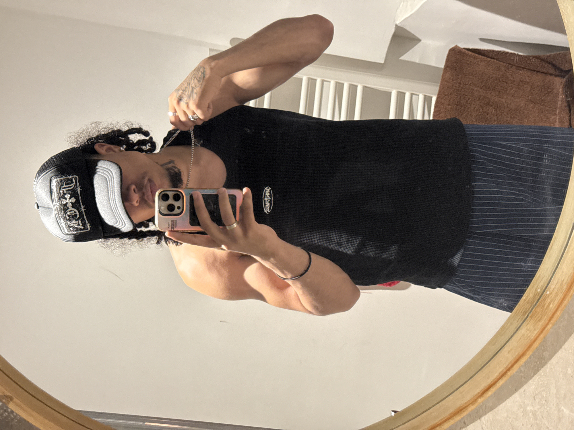 A male individual is taking a selfie in a mirror. He is wearing a black tank top, vertically striped pants, and a black and white baseball cap. He has curly hair and is wearing a silver chain necklace. His left arm is raised, holding the necklace. His right hand holds a pink and gold iPhone. A silver ring is visible on his left ring finger and a silver bracelet is on his left wrist. Tattoos are visible on his left hand and arm. In the background, a white railing, a brown object, and a white ceiling are visible. The mirror has a light wood frame. The location cannot be determined as Massy, France based on this image.