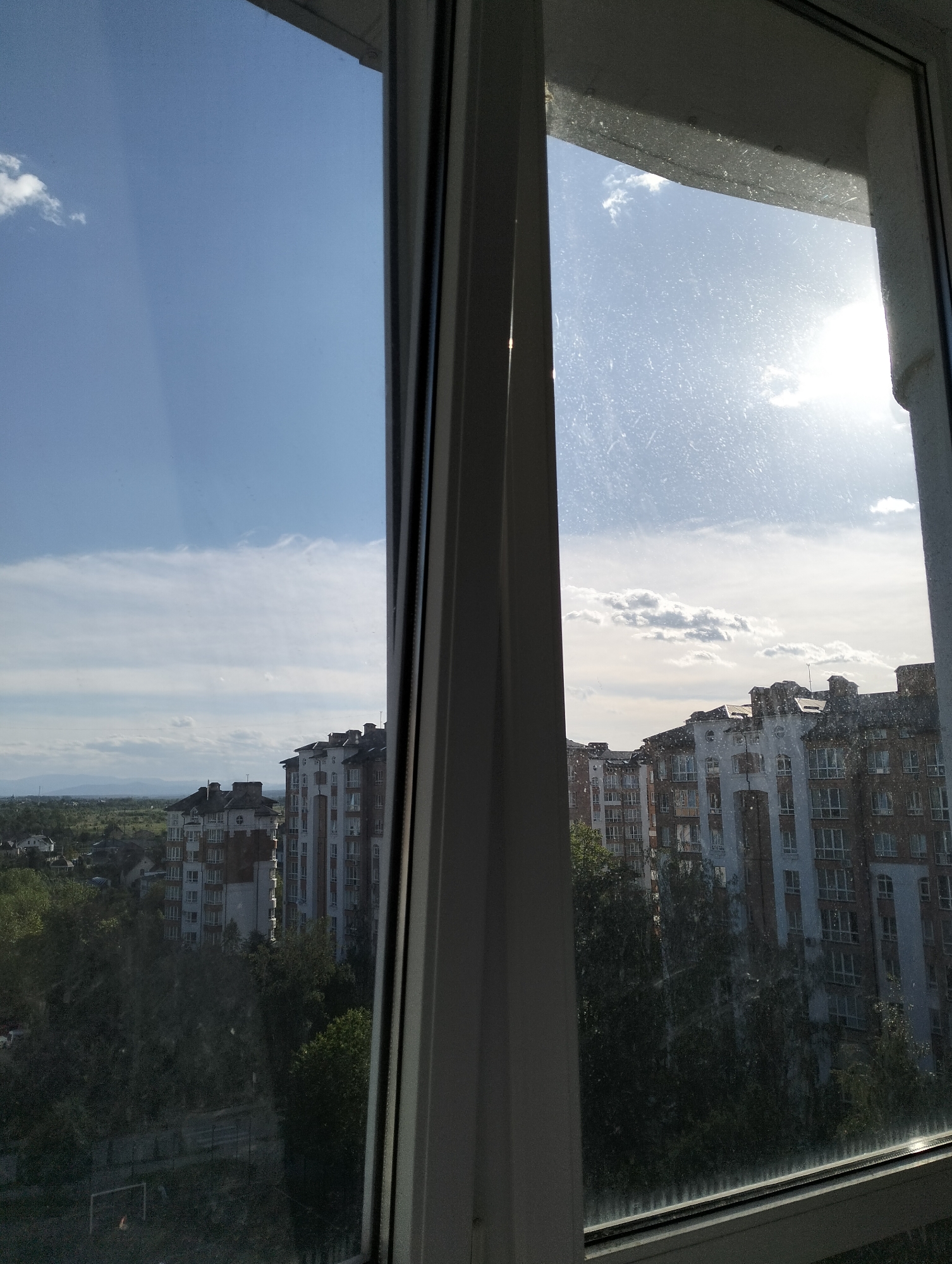 View from a window in Krykhivtsi, Ukraine. The open window frame is white. Outside, there are multiple apartment buildings with light colored facades and dark roofs. Some trees are in front of the buildings. Beyond the buildings, a green area is visible with distant mountains. The sky is blue with scattered clouds and the sun is visible through the window, creating a hazy effect.