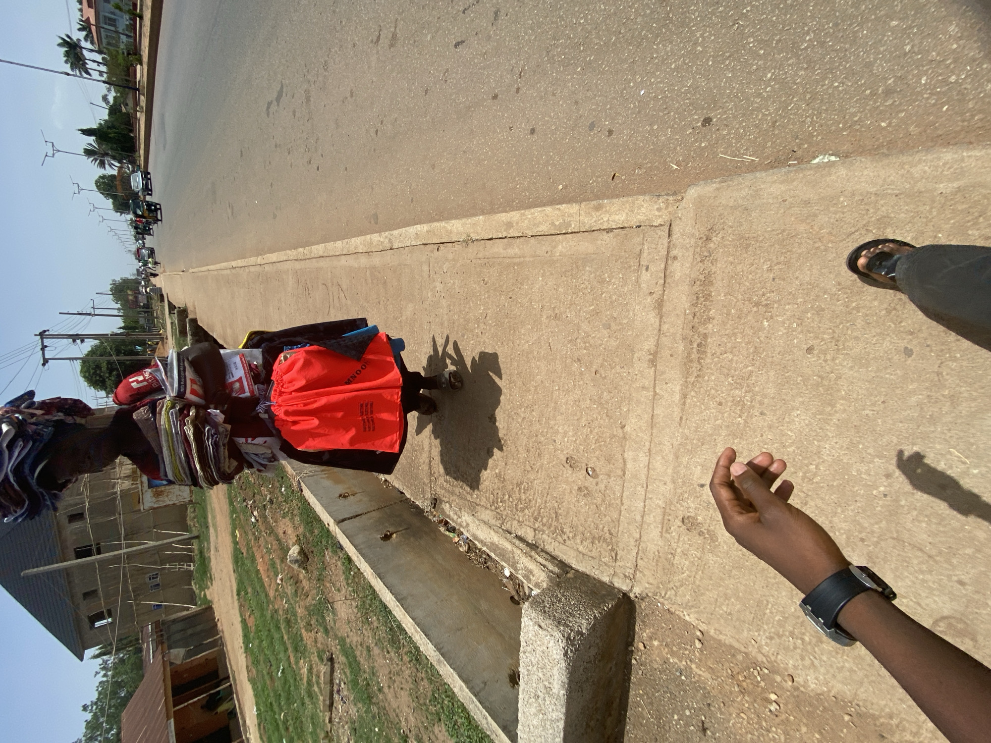 This outdoor image, likely taken in Bukuru, Nigeria, captures a street scene dominated by a vendor and a bustling road under a bright sky. The perspective is from a person walking or standing on a concrete sidewalk, with their right arm (wearing a black watch) and right foot (in black sandals) visible in the foreground on the right.
The primary subject is a person, likely a street vendor, walking on the sidewalk on the left. This individual carries a large and heavy-looking stack of colorful, folded clothes or fabrics on their head, a common hawking method. They also carry several bags, including a prominent red one with white text that partially reads "ONNOO" and features a logo of three horizontal lines, and another red item with the word "EVERYDAY" visible. A white bag with "WITH PERSO" is also seen.
The setting features an asphalt road extending into the distance on the right, with several vehicles, including cars and possibly tricycles, indicating traffic. The sidewalk is made of light-colored concrete, separated from the road by a curb and a concrete drainage gutter that contains some debris. In the background on the left, there are buildings, including one under construction with visible scaffolding, and various trees and utility poles with overhead wires.
The activity depicted is the vendor moving along the sidewalk, presumably hawking their goods. The strong, distinct shadows cast on the ground indicate a sunny day, likely around midday. The clear sky suggests warm weather. The overall scene portrays a typical street environment in a semi-urban or urban area of Nigeria, characterized by local commerce and infrastructure. No explicit emotions are visible, rather it's a snapshot of daily life.