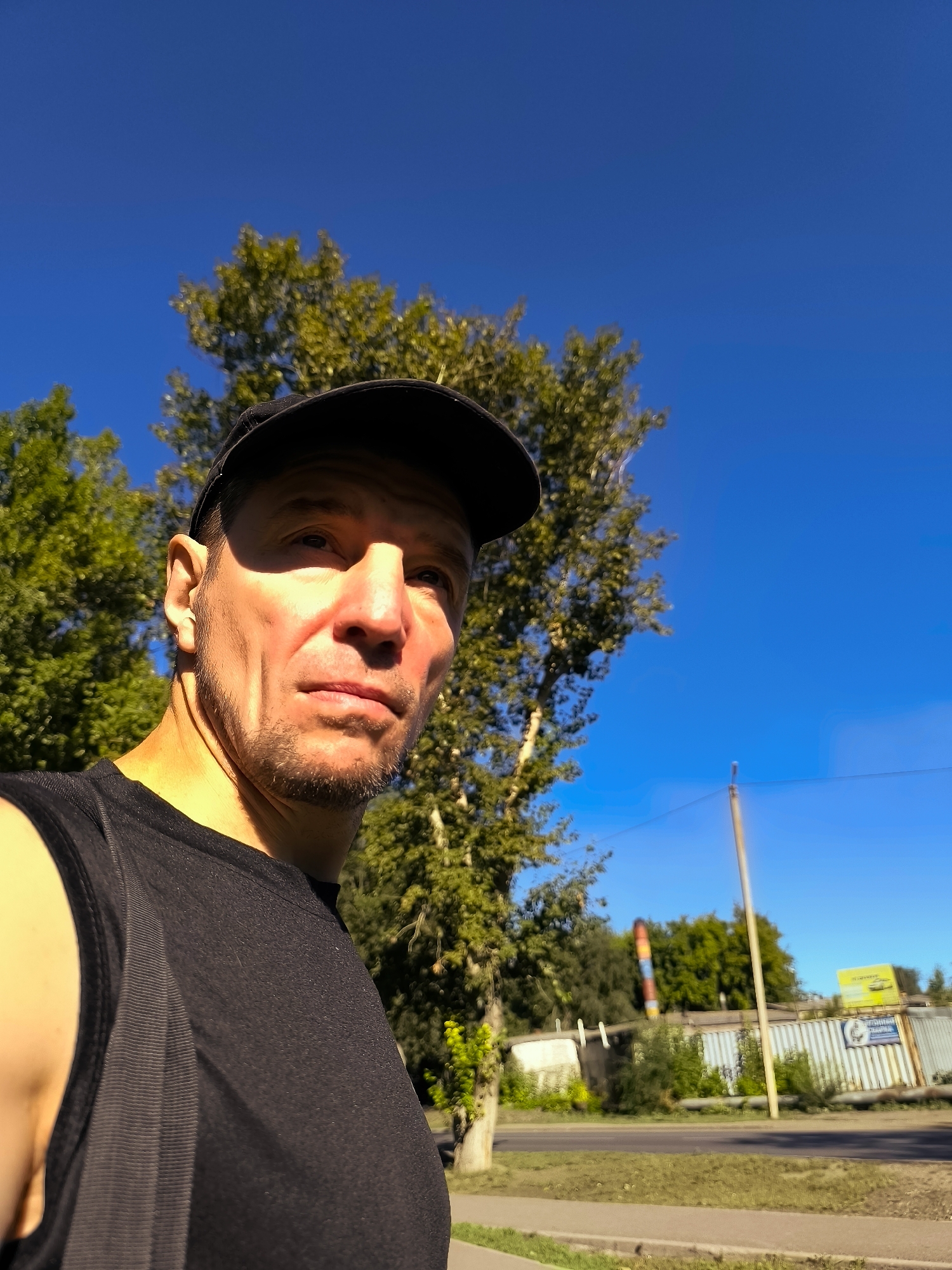 A man in a black sleeveless shirt and a black baseball cap is looking to his right, his face partially illuminated by sunlight. He has short brown hair, a goatee, and is outdoors. Behind him, a tall tree with green leaves is visible against a clear blue sky. In the background, there are buildings, more trees, and a utility pole. The scene appears to be in an urban or suburban area.