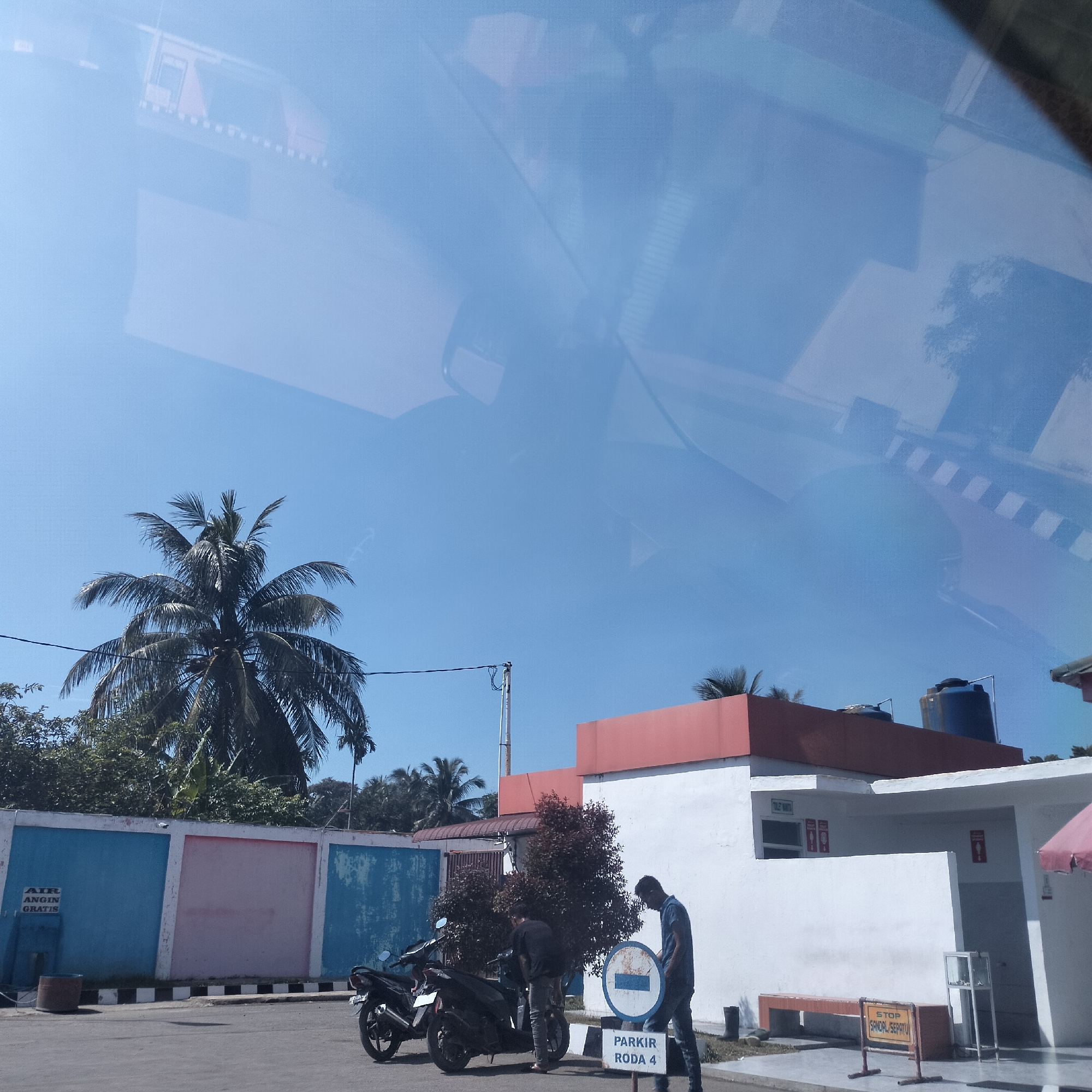 The image presents an outdoor scene captured through a vehicle's windshield on a bright, sunny day, likely at a rest stop or service area in Cotpuuk, Indonesia. The upper portion of the frame shows prominent reflections from the car's interior and external architectural elements.
Two men are visible in the midground. On the right, a man in a dark shirt and jeans stands looking down near a round blue and white sign that reads "PARKIR RODA 4" (Parking for 4-wheel vehicles). To his left, another man in a black shirt is positioned beside a black motorcycle, seemingly attending to it. A second black motorcycle is parked close by.
The background features a white building with a distinctive red roof trim, suggesting public amenities. Several signs are affixed to the building and surrounding structures. On the white building, "TOILET WANITA" (Women's Toilet) and corresponding female icons are visible. A yellow sign below it instructs "STOP SANDAL SEPATU" (Stop Sandals Shoes), indicating an area where footwear should be removed. Further left, a colorful wall with blue and pink segments displays a sign advertising "AIR ANGIN GRATIS" (Free Air).
The setting includes a clear blue sky, casting sharp shadows across the asphalt ground which has zebra-crossing type markings. Lush green foliage, including prominent palm trees, frames the scene. Utility poles and overhead wires are also visible. Water tanks are present on the roof of the white building. The bright conditions indicate it is daytime, likely late morning or early afternoon.
