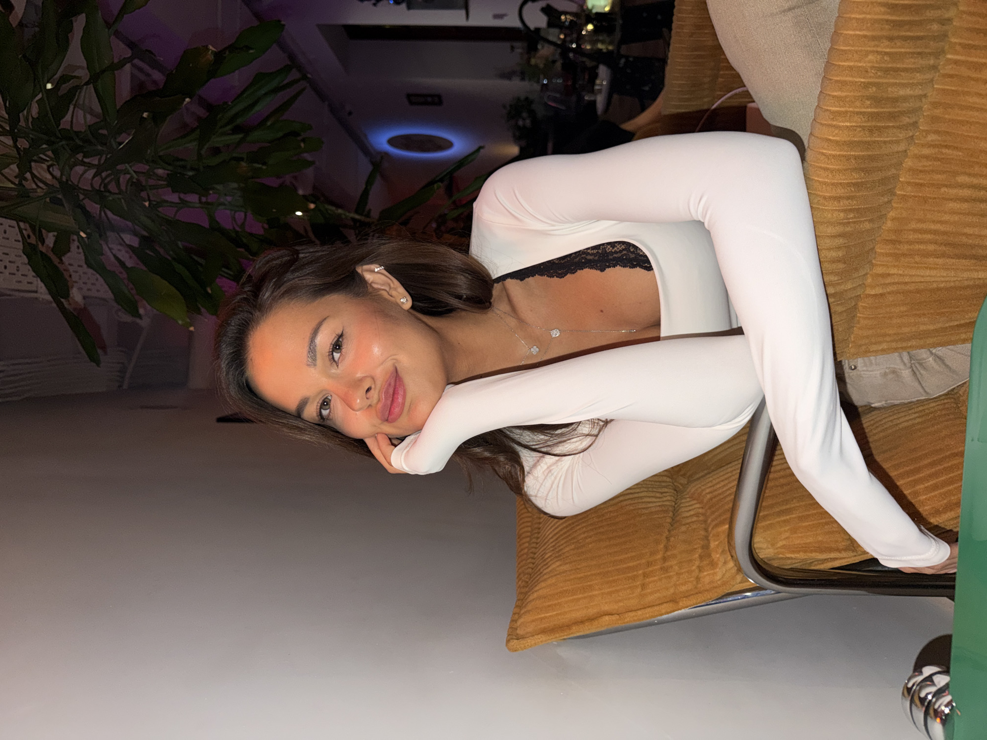 The image captures a young woman posing in an indoor setting, likely a restaurant or lounge, during the evening or night. She is seated on a mustard-yellow, ribbed chair, leaning forward with her chin gently resting on her hand, and looking towards the camera with a soft, subtle smile. She has dark hair, styled down, and wears a white long-sleeved top with a black lace trim visible at the neckline. Delicate jewelry includes small stud earrings and two layered necklaces, one featuring small rectangular pendants and another with clover-shaped elements. In the background, a large green leafy plant is visible, and on the ceiling, a modern light fixture emits a distinct blue glow. The scene conveys a relaxed and comfortable ambiance. No text is visible in the image.