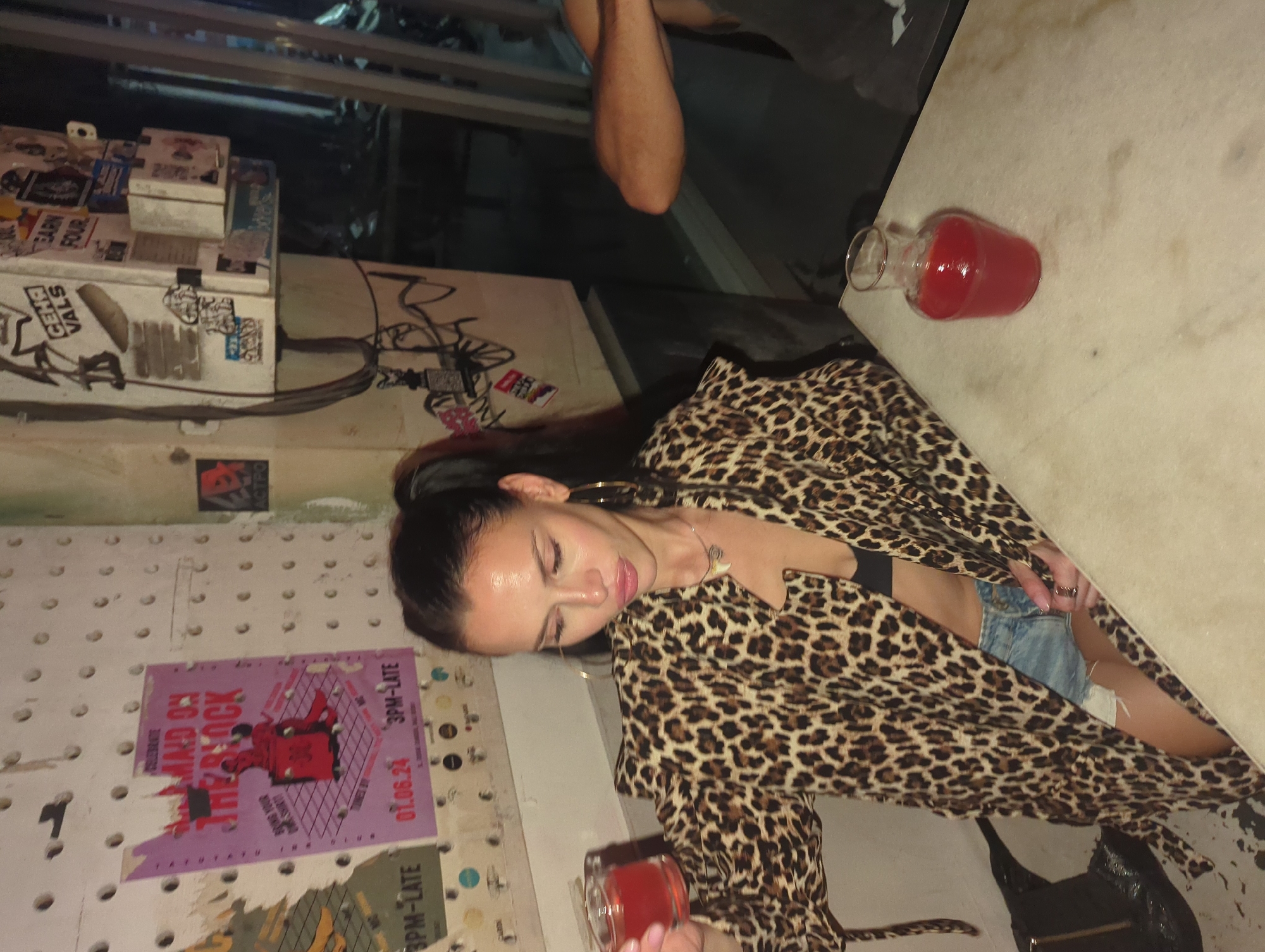 This media file captures a nighttime scene in what appears to be a casual bar or cafe in North Kuta, Indonesia. The main subject is a young woman with dark hair, styled in a bun, wearing a leopard print jacket over a black top and denim shorts. She has large hoop earrings and a necklace. Her eyes are closed or cast downwards, conveying a relaxed or contemplative mood as she holds a glass containing a red drink. Another glass carafe with a red beverage is visible on a light-colored table next to her. A black bag rests on a surface below the table.
In the background, part of a man's arm and leg are visible, suggesting another person is seated or standing nearby. The wall behind the woman is a textured surface, possibly a pegboard, adorned with various posters and stickers. A cluttered ledge with boxes and more stickers is visible on the left side of the image. The dim lighting throughout the scene indicates it is nighttime.
Visible text from posters and stickers includes: "MALDON ON THE BLOCK", "BRING YOUR OWN", "3PM-LATE" (repeated), and the date "01.06.24". Partially visible text reads "TATU TATU INK CLUB". Other fragments of text on stickers include "ACTPO", "СЕНЯ", "VALS", and "EARN FOUR". The overall setting suggests a laid-back, possibly alternative, social venue.