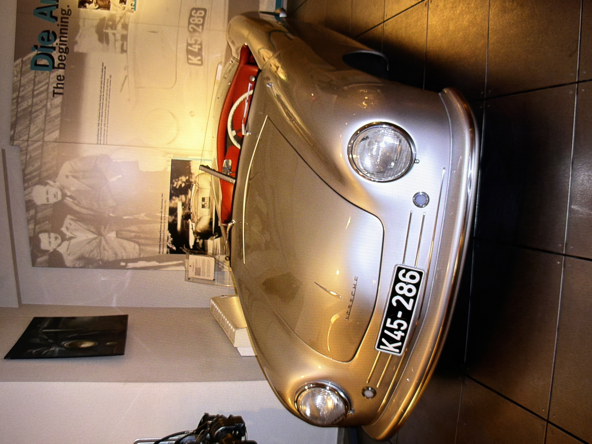 This is a medium shot, eye-level, indoor photograph of a silver Porsche convertible. The car is the primary subject, positioned in the foreground, facing slightly to the left of the camera. Its hood, headlights, and a portion of its interior are visible. The car's license plate displays "K45-286". The interior of the car features red leather seats and a white steering wheel.
In the background, a wall displays a large black and white mural of two men in suits. To the right of the mural, a section of text reads "Die An" followed by "The beginning.". Below the mural, a smaller framed image is visible. To the left of the car, a white radiator is mounted on the wall.
The car is parked on a tiled floor, which appears to be dark brown or black. The lighting in the scene is warm and somewhat dim, suggesting an indoor exhibition or showroom.
Based on the available information and context from the image, this photograph was taken at the Porsche Museum in Kyiv, Ukraine.