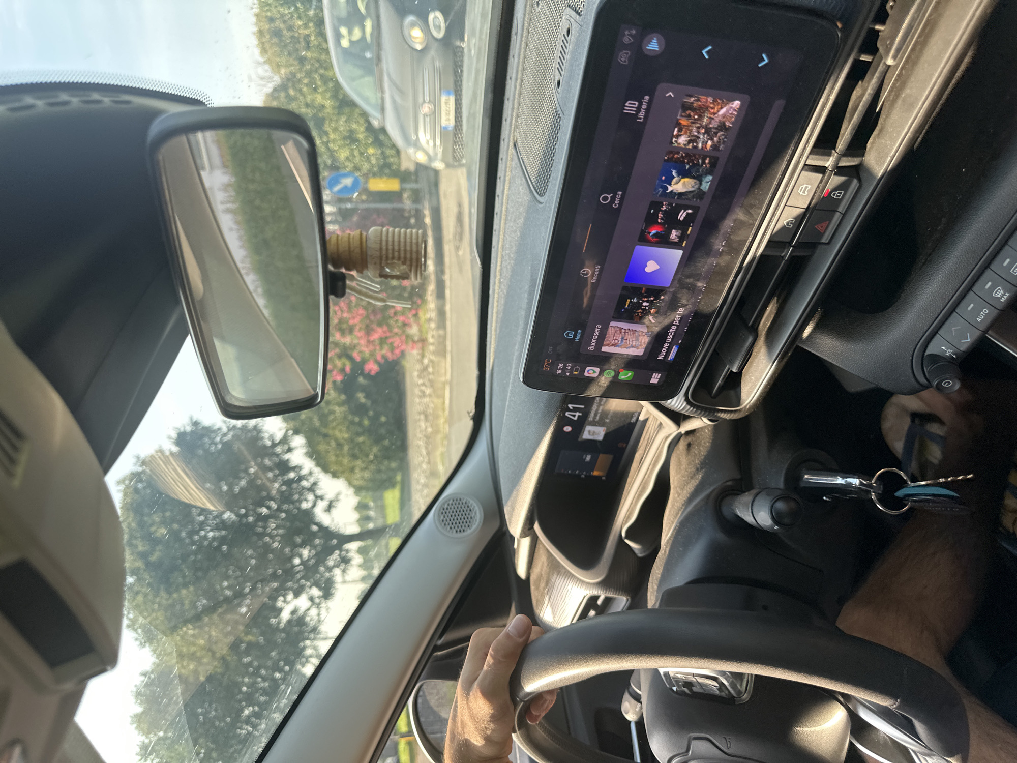 The image captures the interior of a car, looking forward through the windshield. The driver's hand is visible on the steering wheel. The car's dashboard features a large touchscreen display showing a car's infotainment system with various icons and text in Italian. Outside the car, trees and a road are visible, with another car in the distance. The scene appears to be a sunny day. The image was taken in Rudiano, Italy.