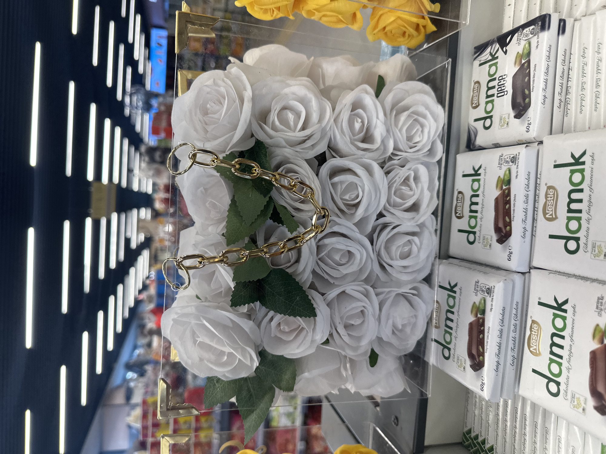 The image shows a display of merchandise inside what appears to be a retail store in Gaziosmanpaşa, Turkey. On the right, there are stacks of "Nestle damak" chocolate bars, including "Antep Fıstıklı Sütlü Çikolata" (Pistachio Milk Chocolate) and "gece" (night) variations. To the left of the chocolates is a clear, rectangular box filled with artificial white roses, adorned with green leaves and a gold chain handle. The roses are arranged densely within the box. The background suggests a store environment with rows of products and shelf lighting.