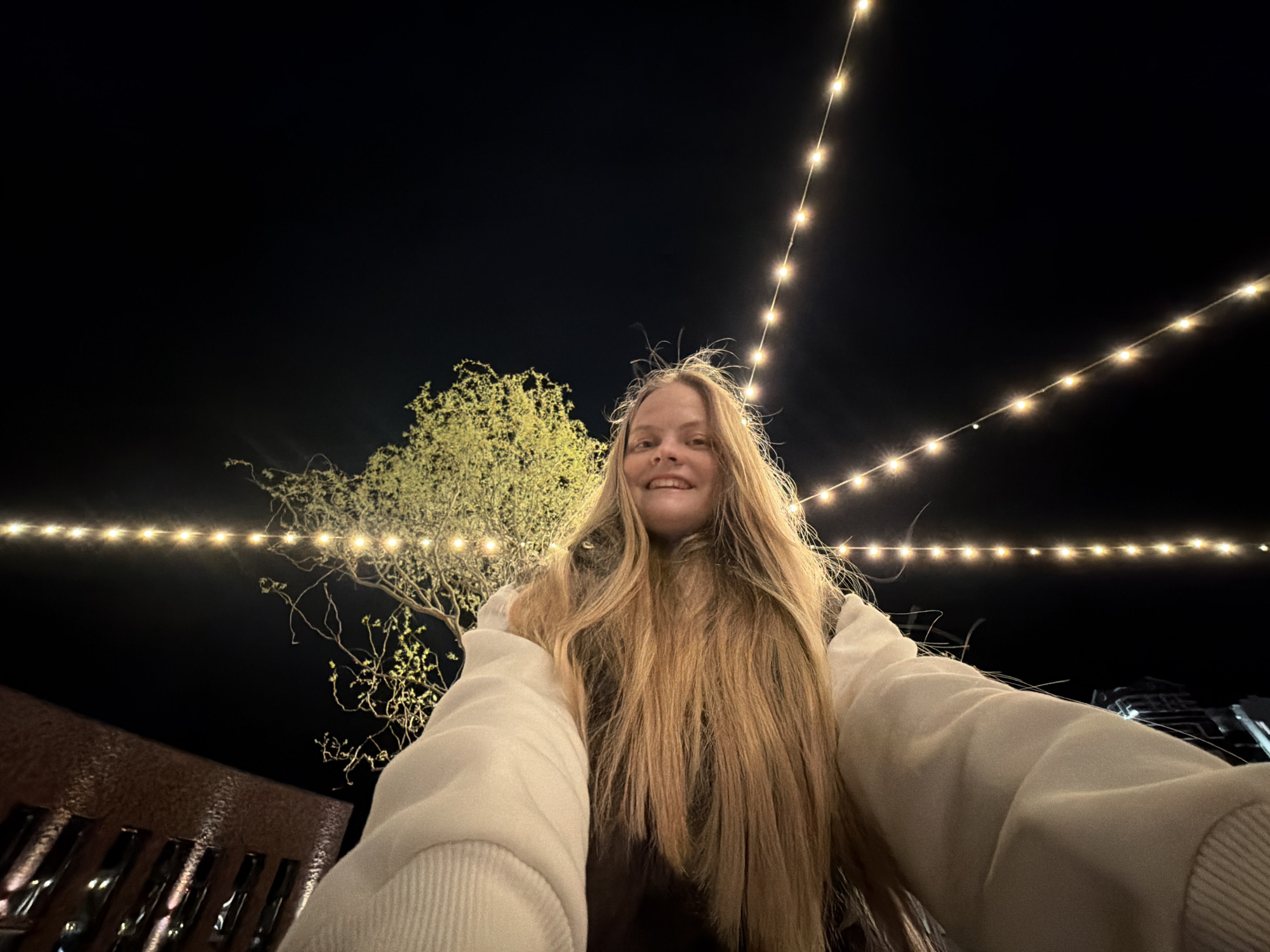 A selfie photograph depicts a young woman at night. She stands beneath strings of lights crisscrossing above and to the sides. Behind her, a tree with budding foliage is visible. The background is a dark sky. A building with a textured facade is partially visible in the lower left corner. She is wearing a light-colored sweater or jacket. The location is not evident from the image and therefore cannot be identified in the city of Izhevsk, Russia.
