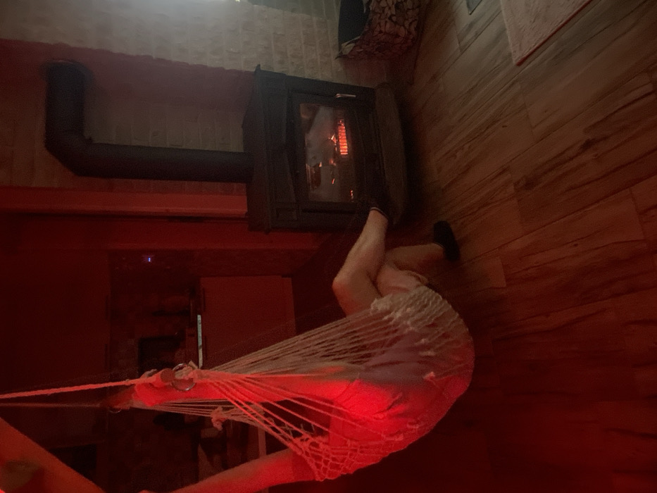 An indoor scene in Palež, Montenegro, depicts a person resting in a hanging macrame chair illuminated by red light. The individual's lower torso, upper legs, and lower legs with dark socks are visible, indicating a seated, relaxed posture with crossed legs resting on the light-colored wooden floor. To the right, a dark, cast-iron wood-burning stove with a visible fire and glowing embers is active. A black stovepipe extends from the top of the stove, bending upwards and to the left. The background features a red-painted wall on the left, with an indistinct counter and a blue light from an electronic device, transitioning to a light-colored brick wall behind the stove on the right. A woven basket and a small rug are partially visible on the floor near the stove. The overall illumination is predominantly red, supplemented by the light from the stove.