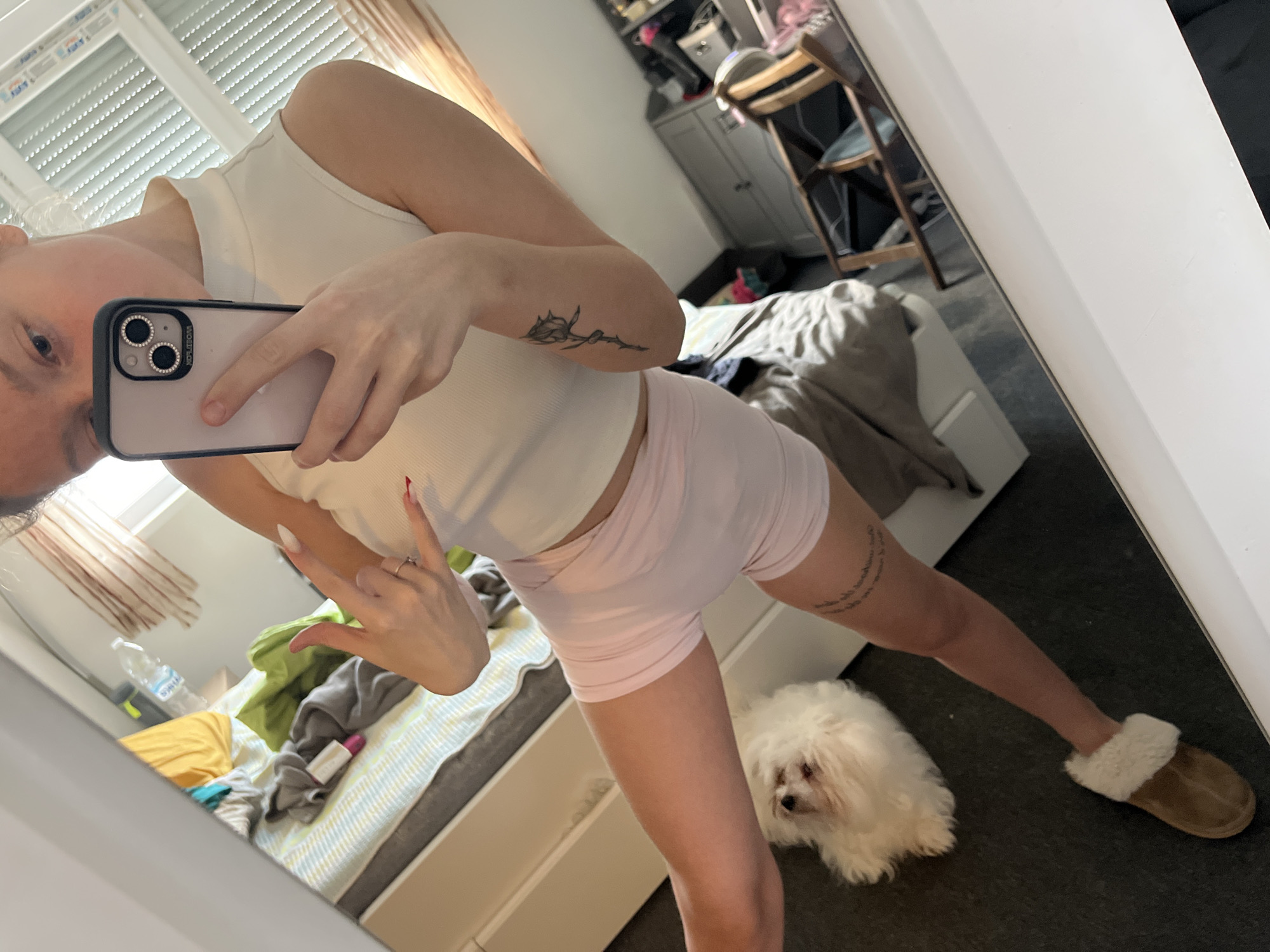A person is taking a selfie in a mirror. They are wearing a white tank top and pink shorts. They are making a peace sign with their left hand. They are holding an iPhone in their right hand. There is a small white dog near their legs, looking towards the camera. They are wearing a brown slipper on their right foot. In the background, there is a bed with striped sheets and various items scattered on it, including clothing and a water bottle. There is a window with blinds behind the bed. Also visible in the background are a grey cabinet, a wooden chair, and items on top of the cabinet. The location is most likely an apartment or home. Varna, Bulgaria is not discernible from the image.
