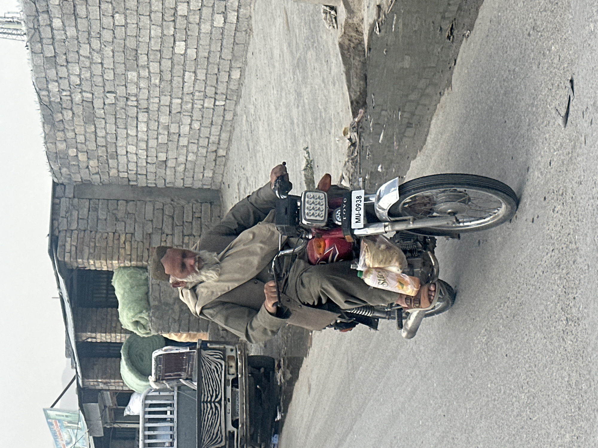 An older man with a white beard, wearing a brown jacket and pants, drives a motorcycle down a street in Quetta City Tehsil, Pakistan. He carries food and other items on the motorcycle, including what appears to be bread. A partially visible building constructed of grey blocks stands to the side of the road, and a vehicle with a zebra stripe pattern is parked nearby. The street is asphalt. The license plate on the motorcycle reads "MU-0938".