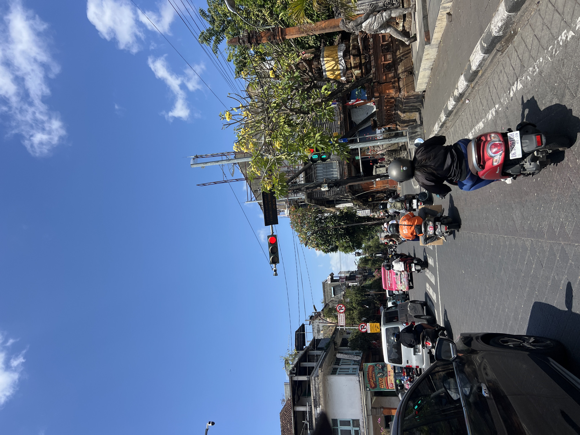 The image captures a street scene in Denpasar, Indonesia, on a sunny day. In the foreground, a black car's side is visible, parked on the left side of the road. Further ahead, a red scooter with a person riding it is moving down the street. Several other motorcycles and a white van are also present in the traffic. Buildings with Balinese architectural elements line the street, adorned with intricate carvings and traditional decorations. A traffic light is visible, showing red, indicating that traffic is stopped. The sky is clear blue with a few scattered clouds. The overall atmosphere suggests a busy urban environment typical of Denpasar.